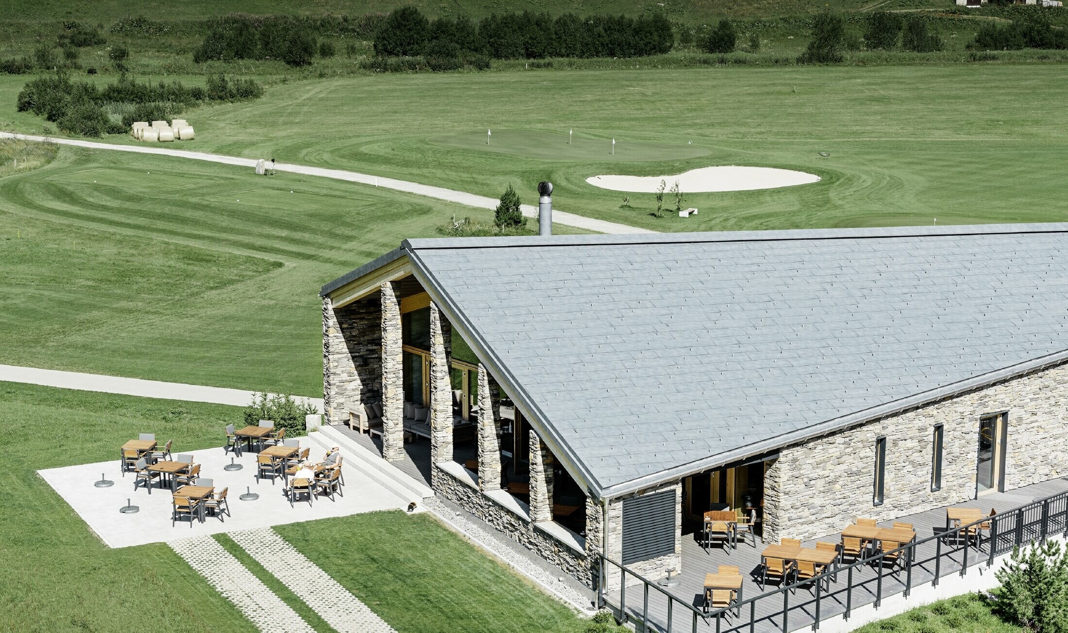 Nowoczesny budynek klubowy na polu golfowym w Andermatt z kamienną elewacją i aluminiowymi panelami dachowymi PREFA FX.12 w kolorze szarym kamiennym