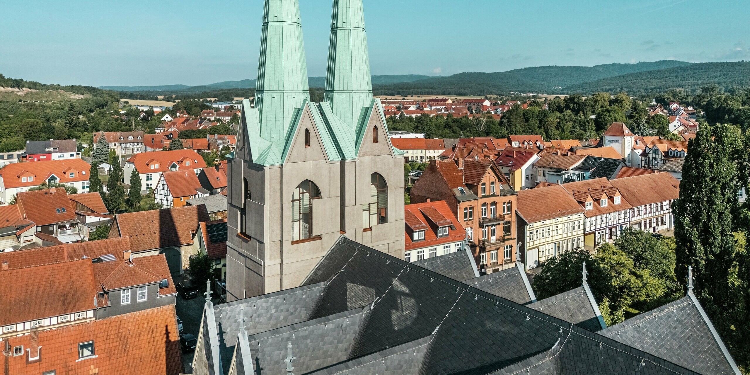 Drohnenaufnahme der neugestalteten Turmspitzen der Stadtkirche St. Johannis in Ellrich, die sich mit PREFALZ in P.10 Patinagrün elegant vom Stadtbild abheben. Die klaren Linien und das moderne Aluminiumdach harmonieren mit der historischen Umgebung und bieten zugleich höchste Wetterbeständigkeit, Langlebigkeit und gestalterische Raffinesse.
