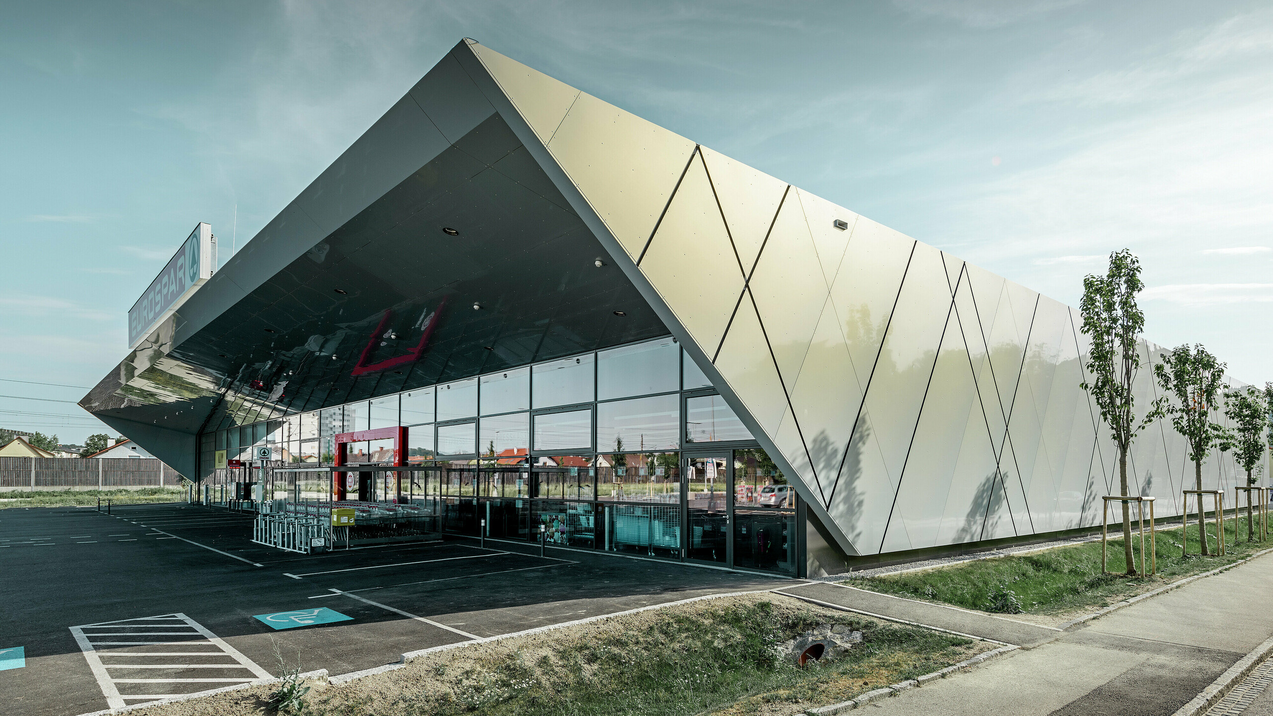 Wejście do nowego sklepu Eurospar w Wels z nowoczesnymi płytami elewacyjnymi PREFA w kolorze silver gold. Aluminiowe płyty kompozytowe połyskują w słońcu różnymi kolorami zależnie od kąta patrzenia.