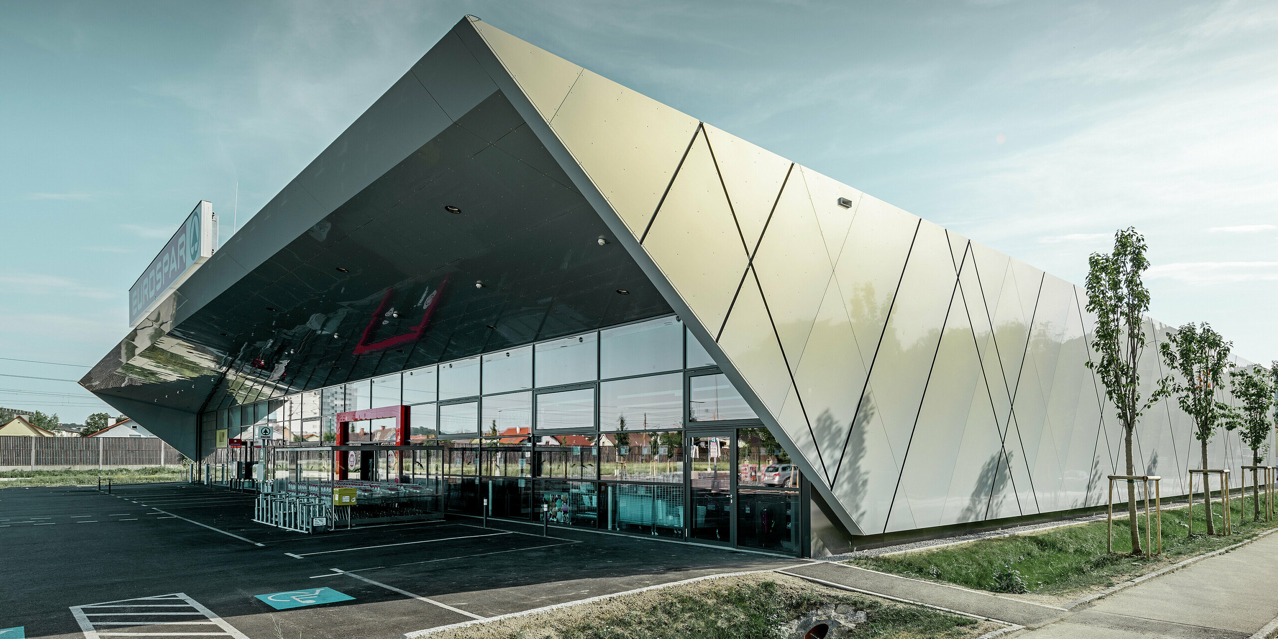 Wejście do nowego sklepu Eurospar w Wels z nowoczesnymi płytami elewacyjnymi PREFA w kolorze silver gold. Aluminiowe płyty kompozytowe połyskują w słońcu różnymi kolorami zależnie od kąta patrzenia.
