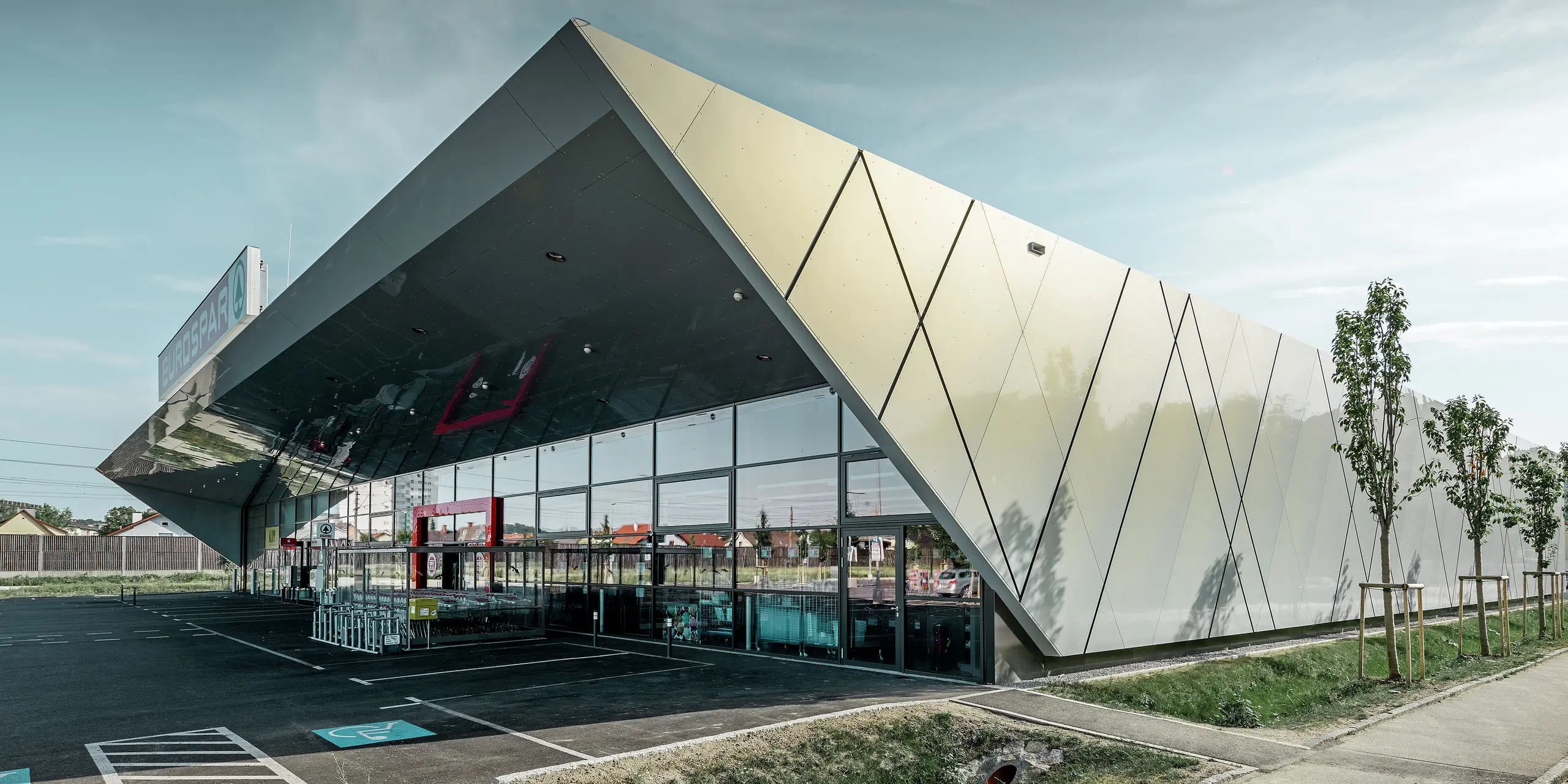 Wejście do nowego sklepu Eurospar w Wels z nowoczesnymi płytami elewacyjnymi PREFA w kolorze silver gold. Aluminiowe płyty kompozytowe połyskują w słońcu różnymi kolorami zależnie od kąta patrzenia.