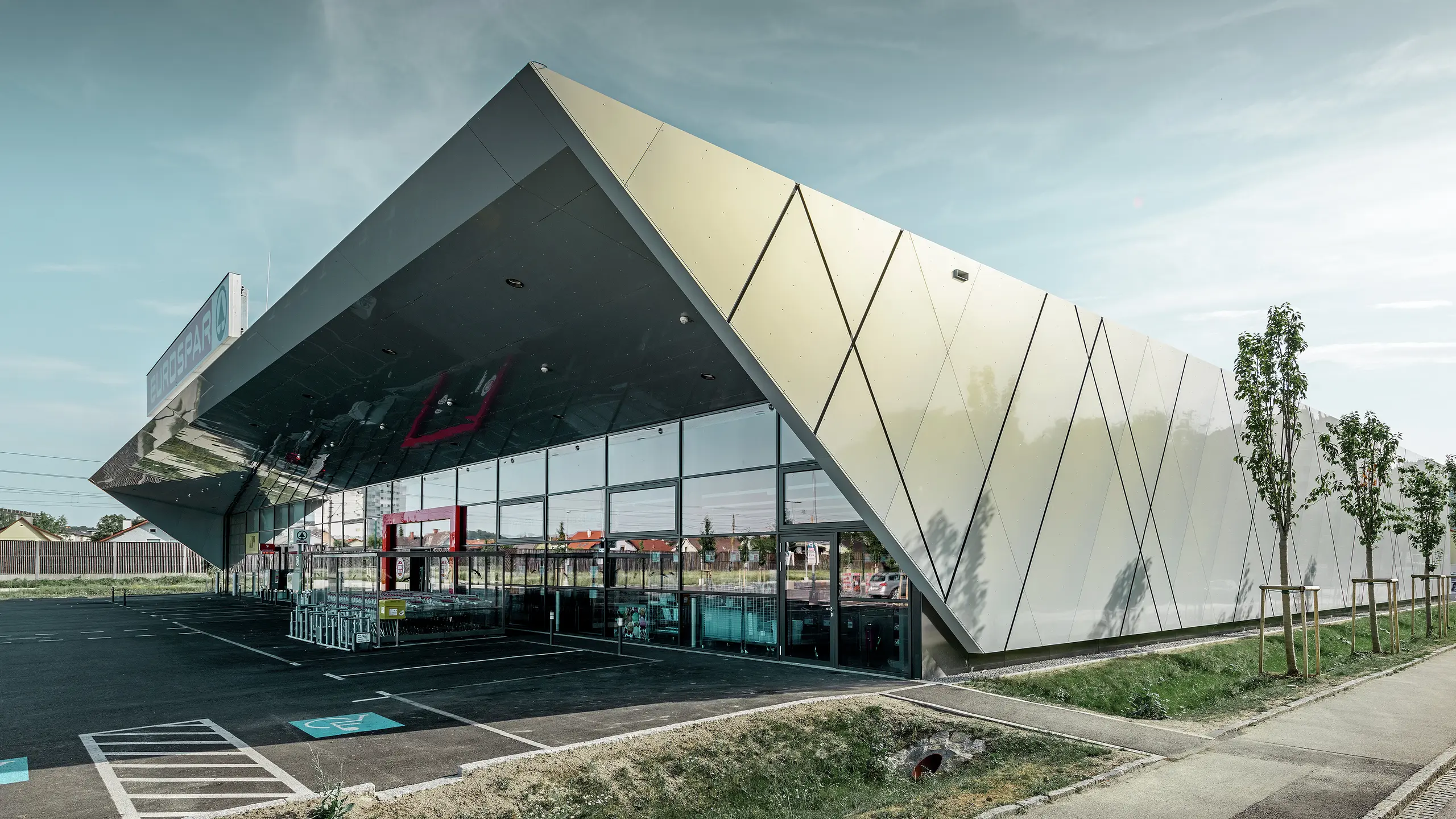Wejście do nowego sklepu Eurospar w Wels z nowoczesnymi płytami elewacyjnymi PREFA w kolorze silver gold. Aluminiowe płyty kompozytowe połyskują w słońcu różnymi kolorami zależnie od kąta patrzenia.