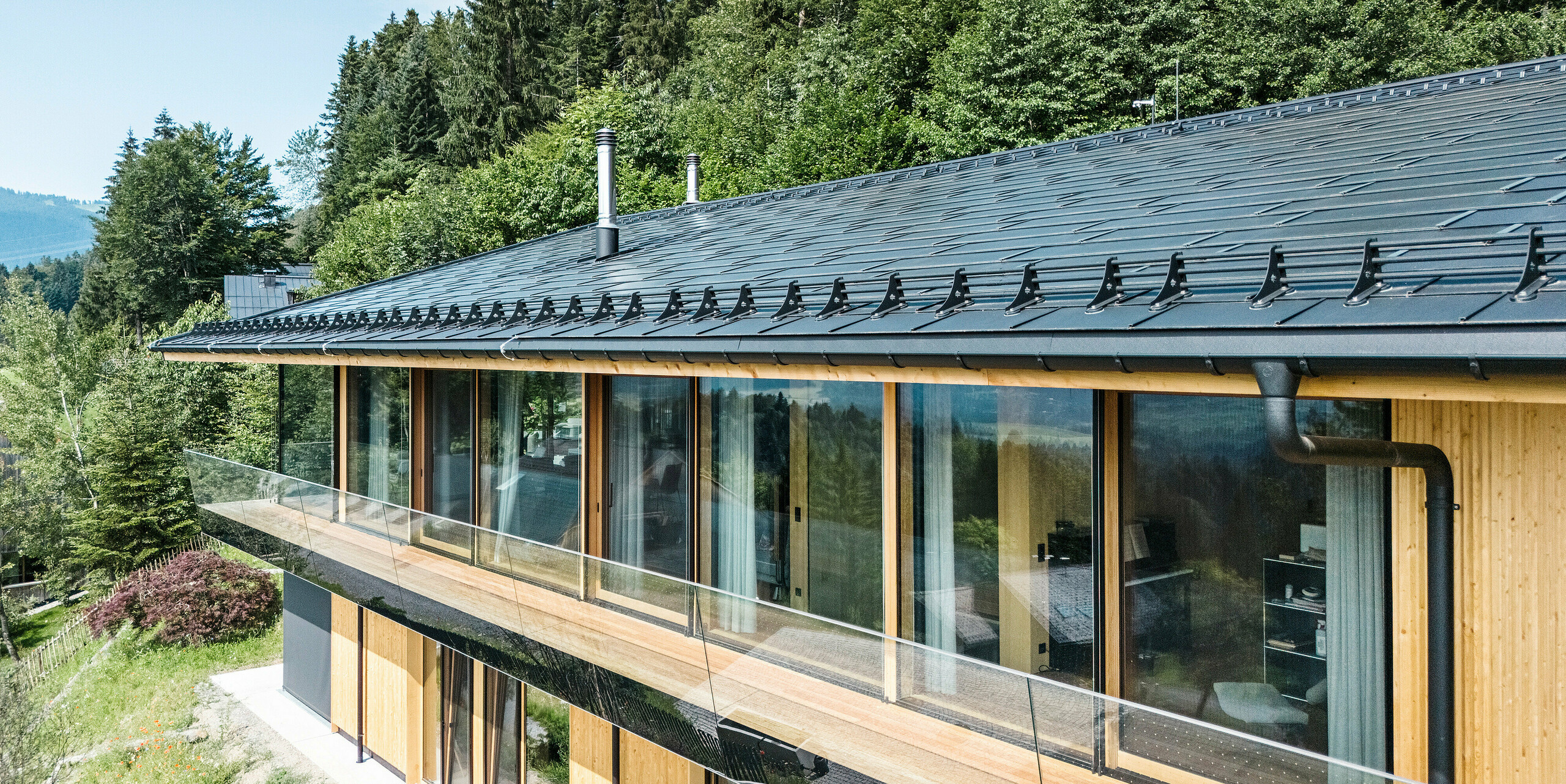 Seitenansicht eines architektonisch anspruchsvollen Einfamilienhauses in Egg, Vorarlberg, mit PREFA Solardachplatten und Dachplatten R.16 in P.10 Schwarz. Die Aluminium-Dacheindeckung harmoniert perfekt mit der modernen Glasfassade und der natürlichen Umgebung. Zu sehen ist auch das effektive Schneefangsystem sowie die nahtlos integrierte PREFA Dachentwässerung mit Hängerinne und Ablaufrohr.