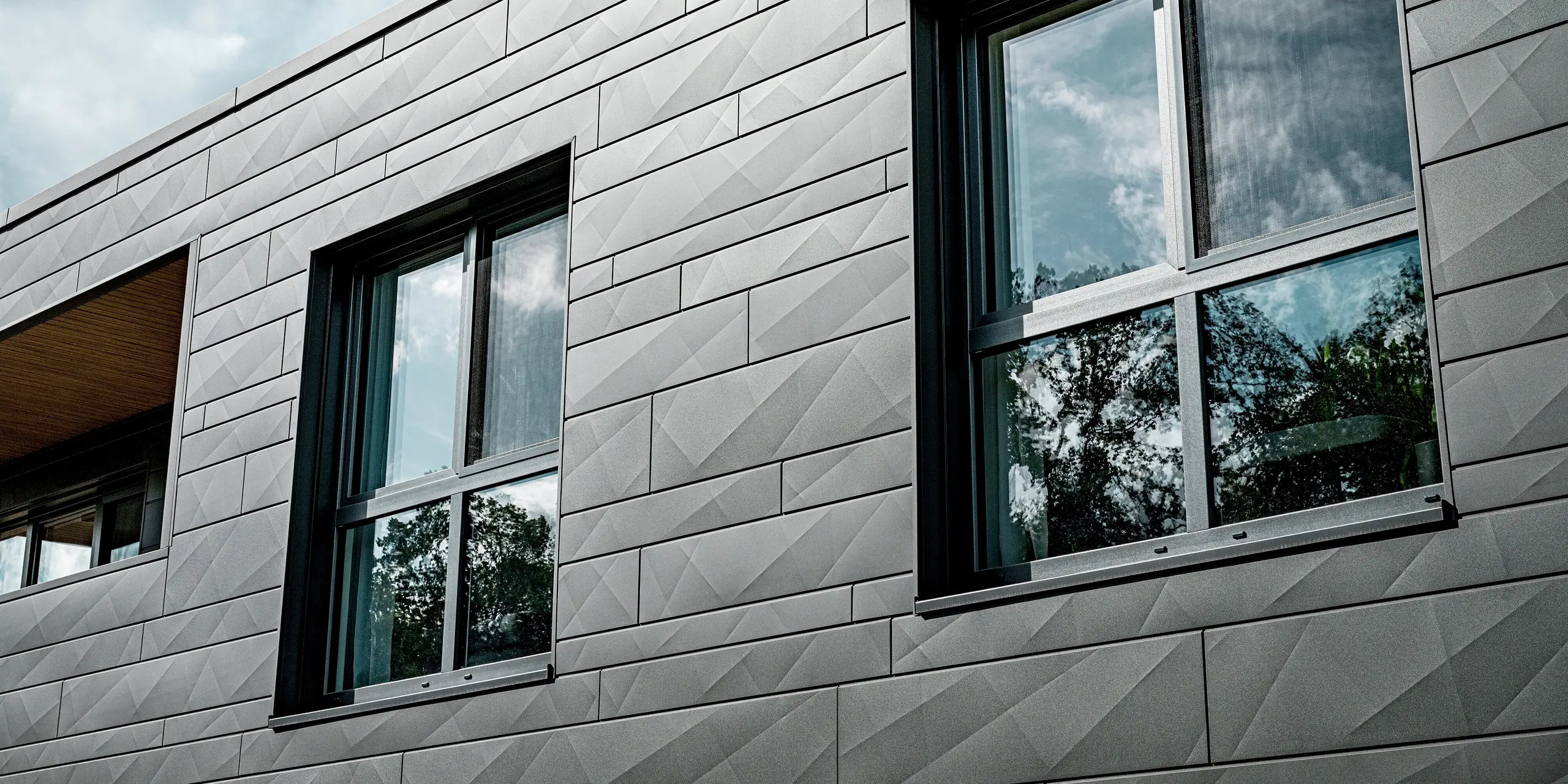 Nahaufnahme der PREFA Siding.X Aluminiumfassade in P.10 Dunkelgrau an einem modernen Einfamilienhaus in Heidenheim. Die unterschiedlich breiten Paneele mit markanten Kantungen erzeugen ein lebendiges, geometrisches Lichtspiel. Zwei große Fenster spiegeln Himmel und Baumkronen und betonen die klare, reduzierte Architektur. Der präzise Abschluss der Paneele zeigt die hohe Verarbeitungsqualität und die langlebige, witterungsbeständige Aluminiumoberfläche, die das Gebäude dauerhaft schützt und dessen moderne Optik unterstreicht.