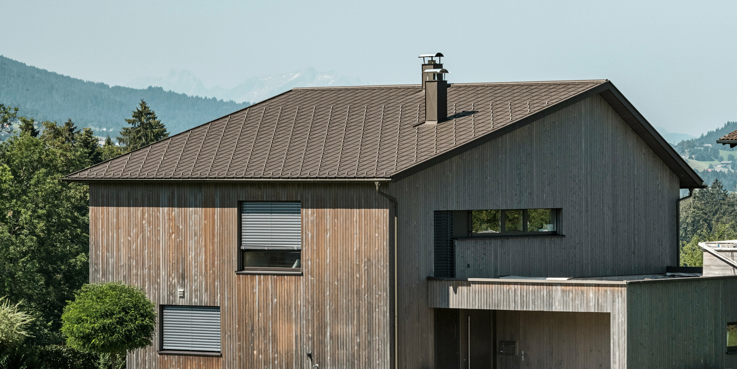 Seitenansicht eines zeitgenössischen Einfamilienhauses in Krumbach mit einer Kombination aus einer natürlichen Holzfassade und einem langlebigen PREFA Aluminiumdach in P.10 Braun. Die harmonische Farbgebung zeigt die elegante Integration von modernem Design und traditionellen Materialien, wobei die PREFA Dachplatten für eine ästhetische und funktionale Dachgestaltung sorgen.