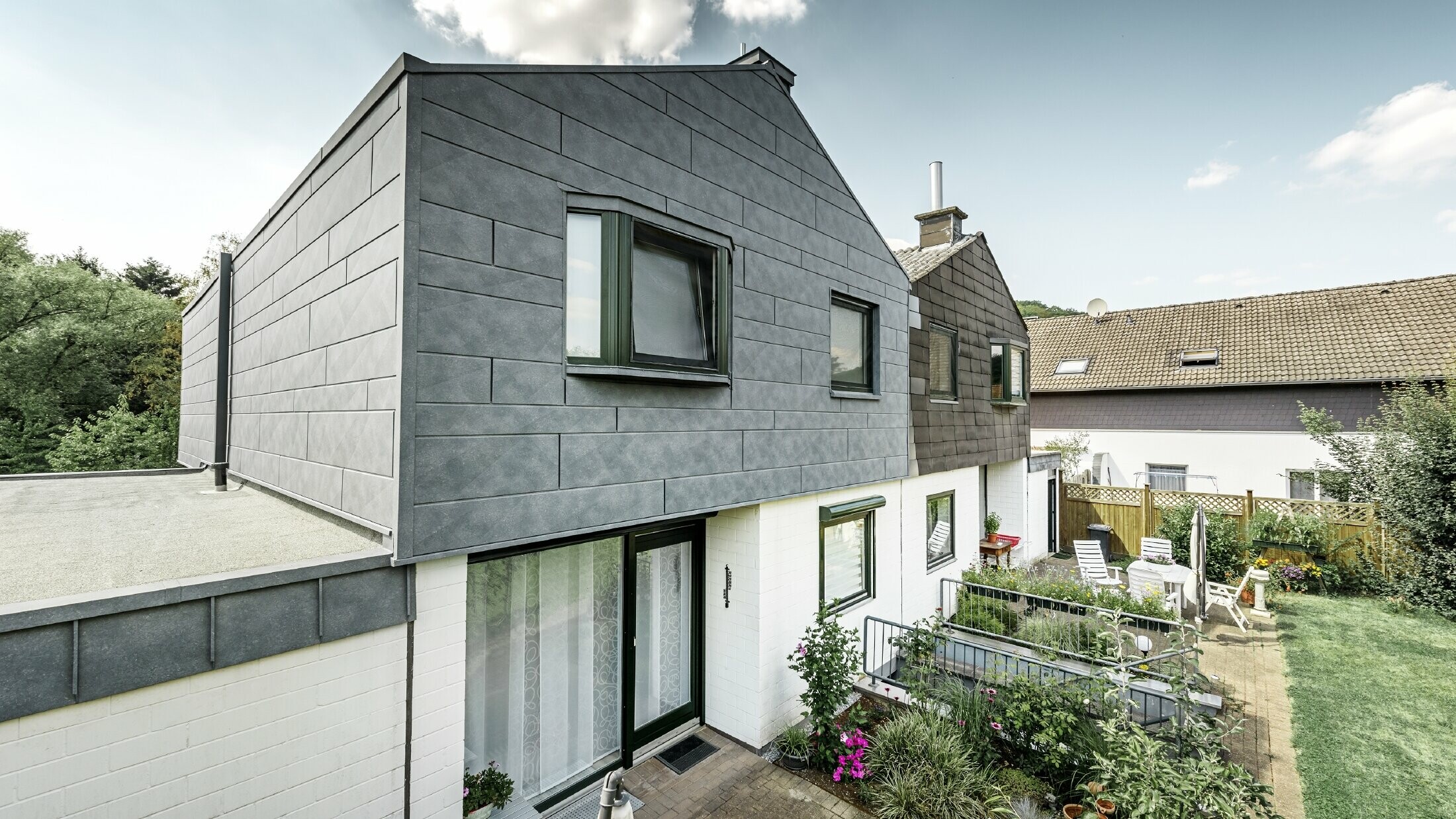 Renowacja górnej części połówki bliźniaka została przeprowadzona przy użyciu elementu elewacyjnego PREFA Siding.X w kolorze szarym kamiennym P.10. Obok dla porównania połówka domu przed renowacją.