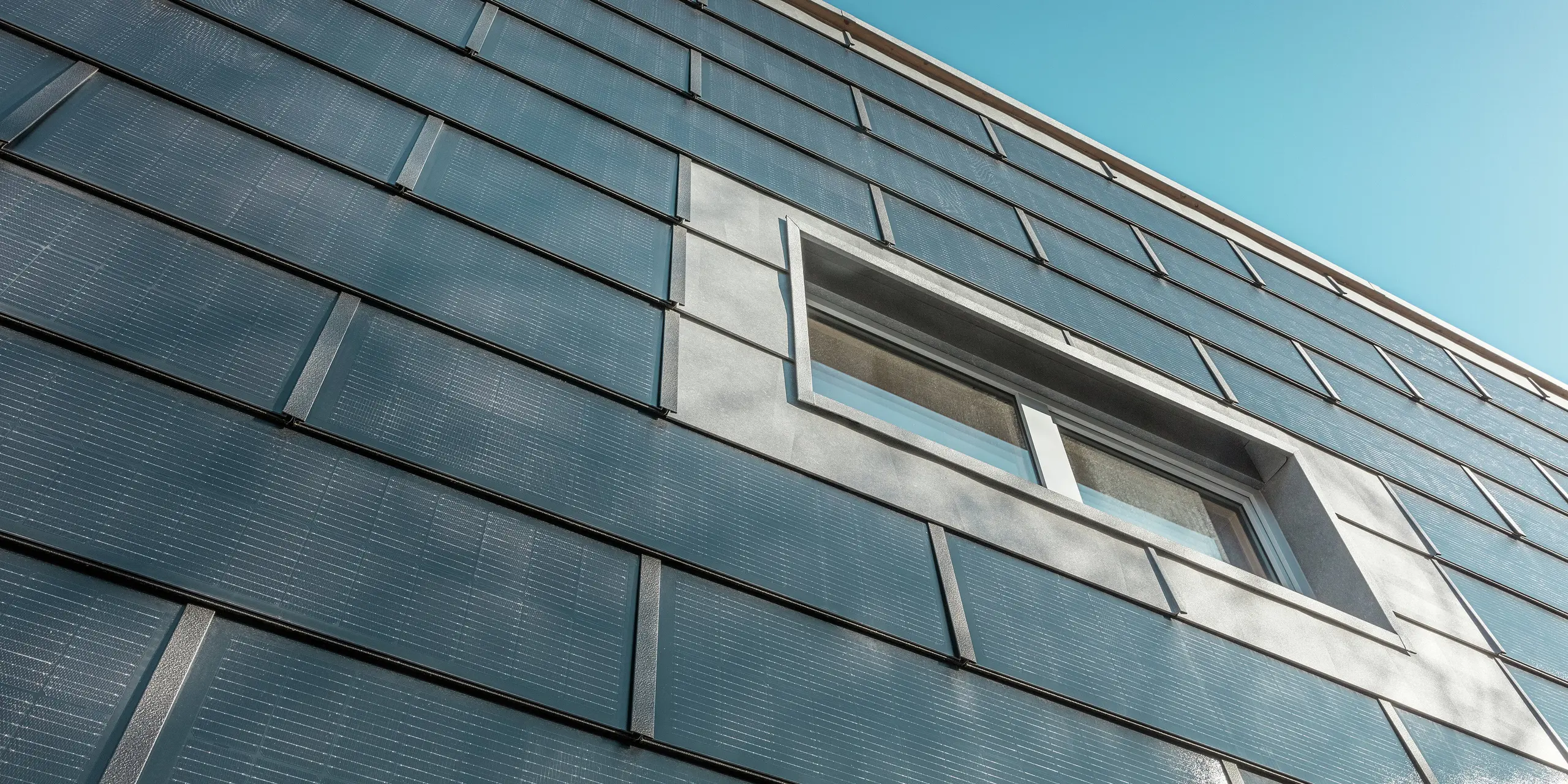 Die Fassade der RÜMA-Blechwerkstatt in Vorarlberg, aus einem bestimmten Winkel fotografiert, ist mit PREFA-Solardachziegeln verkleidet. Graue Aluminiumelemente und integrierte Photovoltaikmodule rahmen das Fenster ein. Im Hintergrund ist der klare blaue Himmel zu sehen.