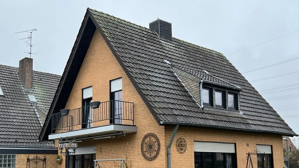 Dom jednorodzinny z żółtej cegły klinkierowej z stromym dachem dwuspadowym i ciemnym pokryciem dachowym przed renowacją dachu; od strony ogrodu z balkonem, lukarną, tarasem z meblami ogrodowymi i trawnikiem na pierwszym planie.