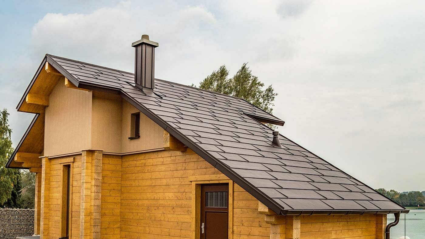 Dom nad jeziorem z panelem dachowym PREFA FX.12 w kolorze brązowym orzechowym, z okładziną elewacji w kolorze brązowym piaskowym i w kolorze drewna.