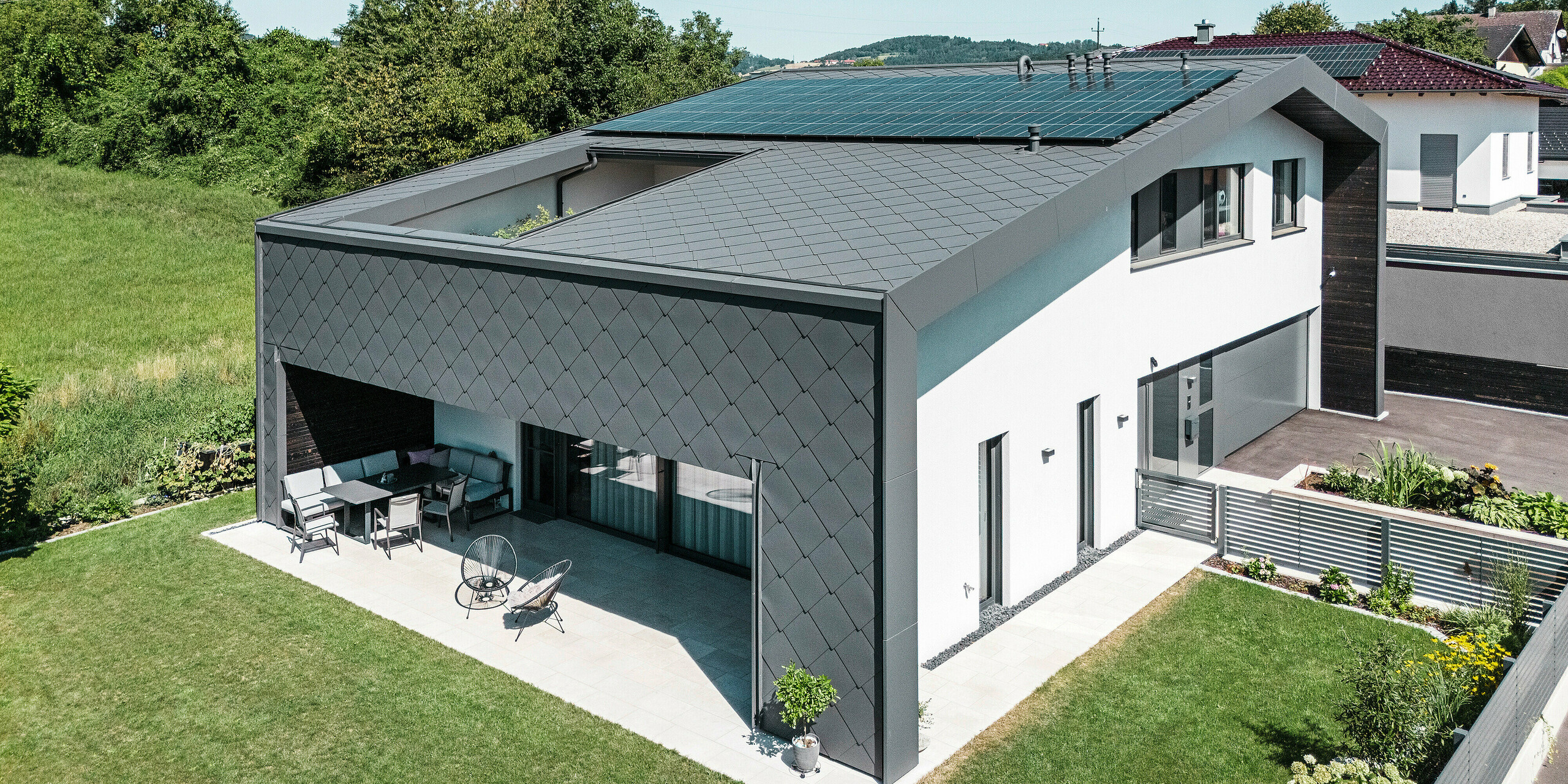 Modernes Einfamilienhaus in Aschach mit eleganter Aluminiumbekleidung aus dunkelgrauen PREFA Dach- und Wandrauten 44 × 44 in der hochwertigen Beschichtung P.10. Das Gebäude zeichnet sich durch eine großzügige, überdachte Terrasse aus, die fließend in den gepflegten Garten übergeht. Eine Photovoltaikanlage auf dem Dach sorgt für nachhaltige Energiegewinnung. Die Kombination aus moderner Architektur, witterungsbeständiger Aluminiumhülle und schlichter weißer Putzfassade verleiht dem Haus ein stilvolles und zeitgemäßes Erscheinungsbild.