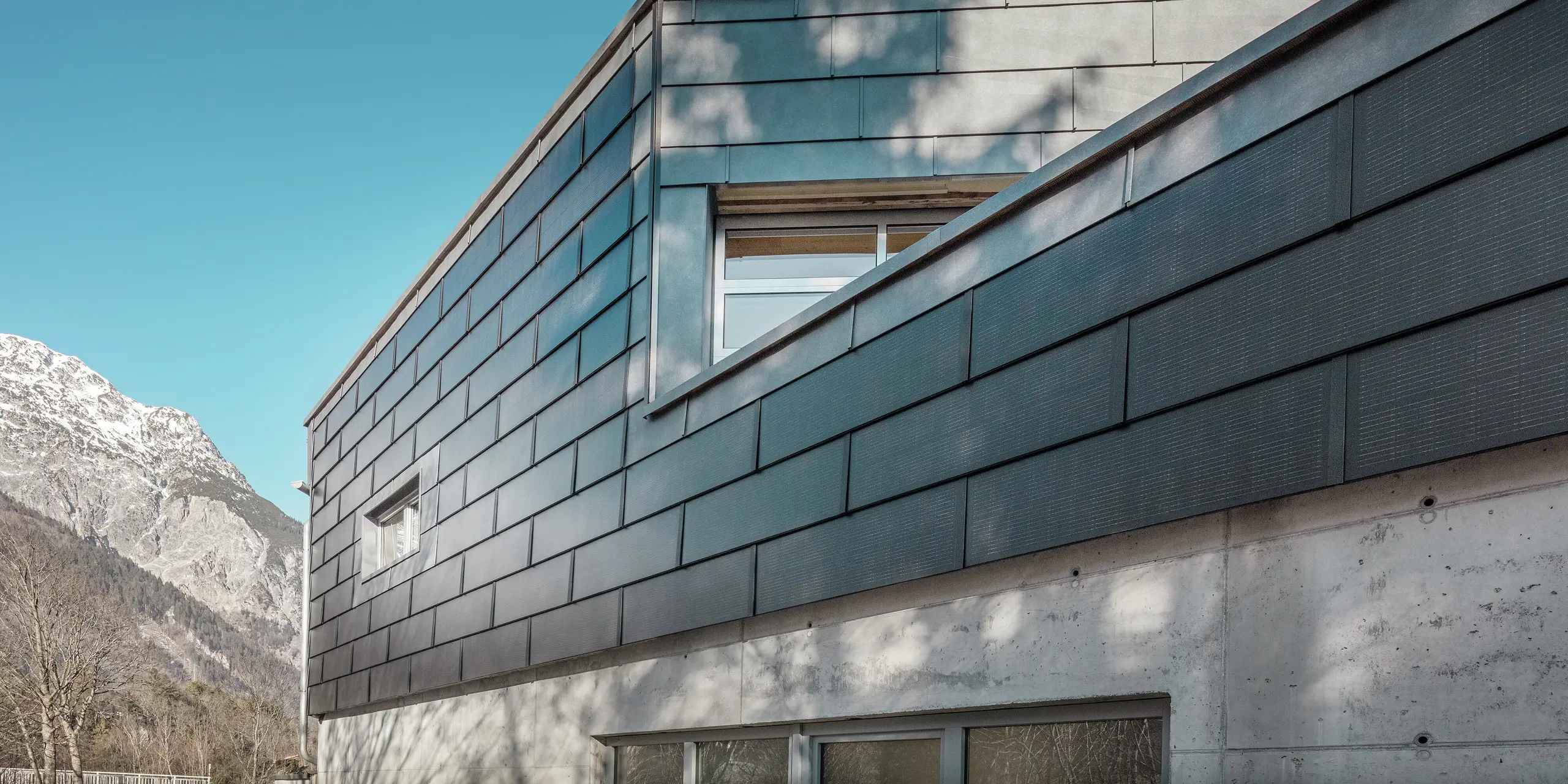 Die Fassade des Bürogebäudes der RÜMA Blechwerkstatt in Vorarlberg mit innovativen PREFA Solardachpaneelen und Sichtbeton, umgeben von Bergen unter klarem blauem Himmel, mit Baumschatten an der Wand.