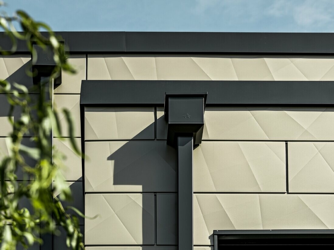 Nowoczesny dom jednorodzinny z płaskim dachem i wąskimi oknami. Elewacja jest pokryta panelami PREFA Siding.X z efektem 3D, w kolorze brązowym. Połączenia wykonano w kolorze antracytowym. Do odwodnienia dachu zastosowano rurę kwadratową z dopasowanym koszem zlewiskowym w kolorze antracytowym.