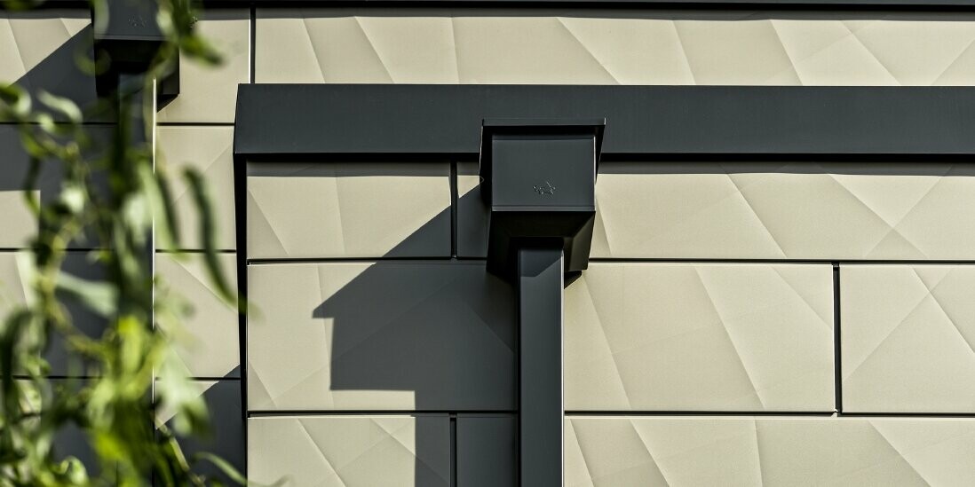 Nowoczesny dom jednorodzinny z płaskim dachem i wąskimi oknami. Elewacja jest pokryta panelami PREFA Siding.X z efektem 3D, w kolorze brązowym. Połączenia wykonano w kolorze antracytowym. Do odwodnienia dachu zastosowano rurę kwadratową z dopasowanym koszem zlewiskowym w kolorze antracytowym.