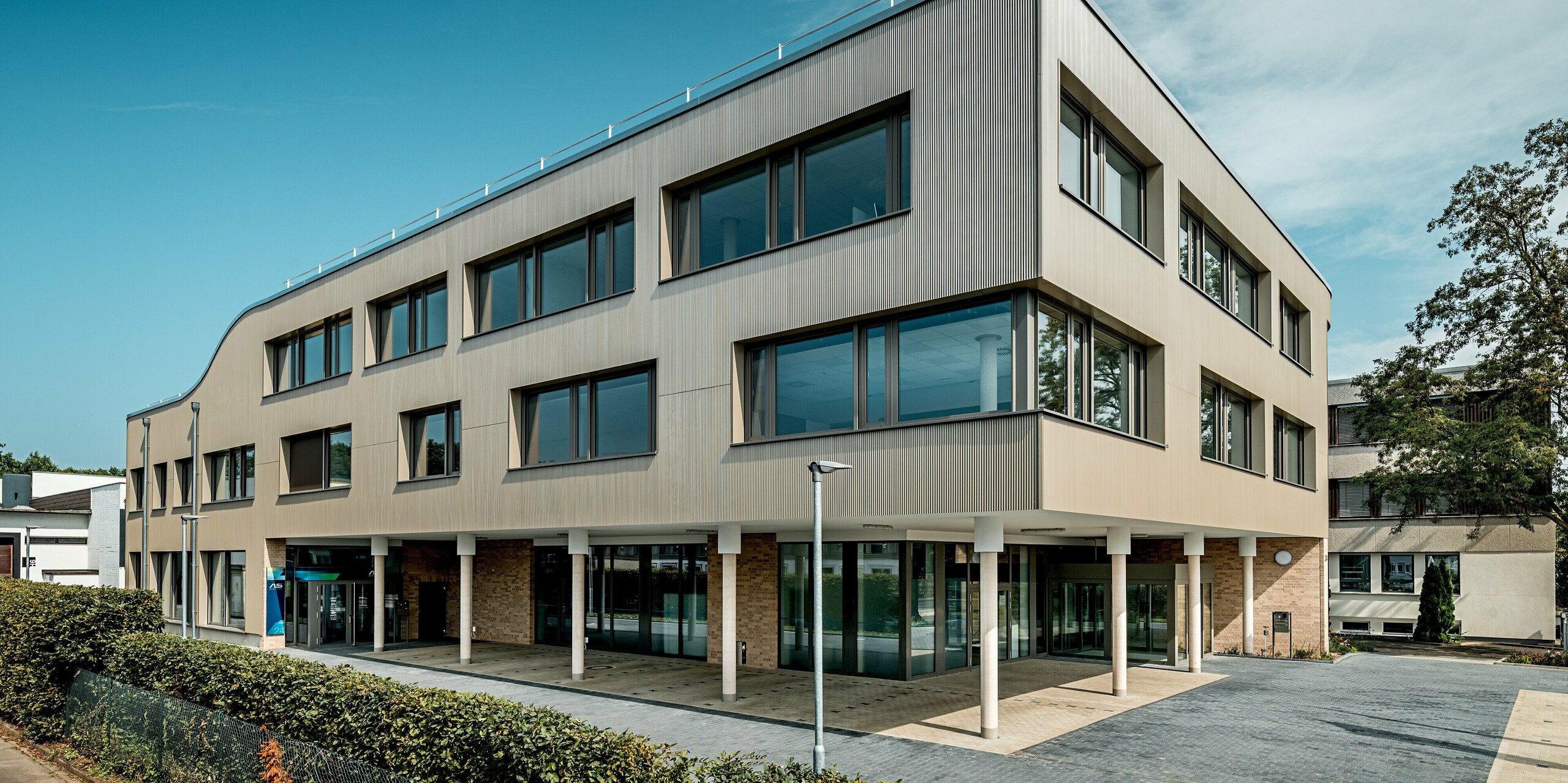 Das Bild zeigt den Eingangsbereich des modernen Festo-Gebäudes in Langenhagen, das durch eine markante, geschwungene Fassade besticht. Die Fassade ist mit PREFA Strangpressprofilen in Zackenform verkleidet, die eine interessante Textur und Tiefe erzeugen. Die klaren Fensterflächen und die glatte, elegante Struktur der Aluminium Verbundplatten sorgen für eine harmonische Kombination aus Funktionalität und Design. Besonders hervorzuheben ist die sanft abgerundete, obere Kante des Gebäudes, die dem gesamten Bauwerk eine moderne, fließende Ausstrahlung verleiht. 