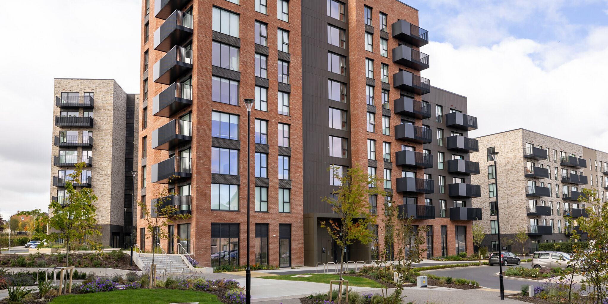 Frontansicht der Citywest Apartments in Dublin. Das Wohngebäude kombiniert eine traditionelle Ziegelfassade mit modernem Design aus vertikal verlegten PREFA Sidings in P.10 Braun. Großzügige Balkone und großzügige Fensterflächen unterstreichen die moderne Architektur. Auf ca. 700 m² bietet die witterungsbeständige Aluminiumfassade eine stilvolle und langlebige Lösung, die sich harmonisch in die städtebaulich gestaltete Umgebung mit gepflegter Grünanlage einfügt.