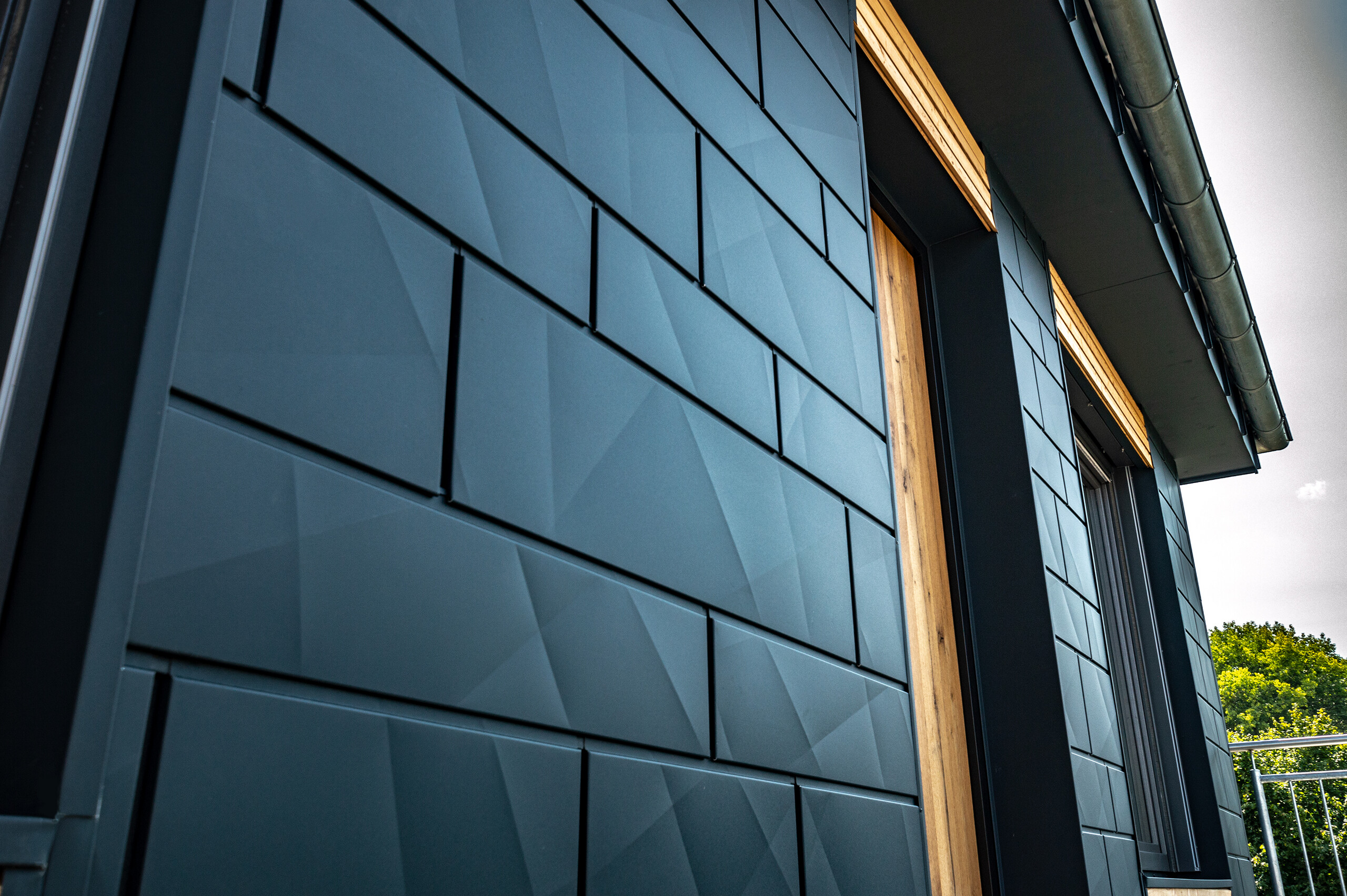 Elewacja z panelami PREFA Siding.X w kolorze antracyt, z drzwiami balkonowymi i dużymi przeszkleniami, połączona z elewacją drewnianą.