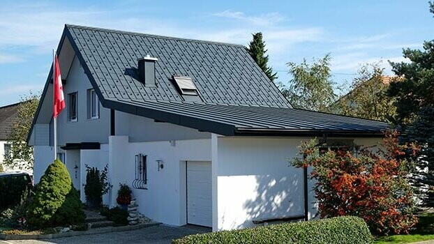 Zmodernizowany dom z dachem szczytowym i przyłączonym garażem. Dach został pokryty płytą dachową PREFA, a garaż blachą Prefalz w kolorze antracytu. Przed domem znajduje się maszt na flagę z flagą Szwajcarii.