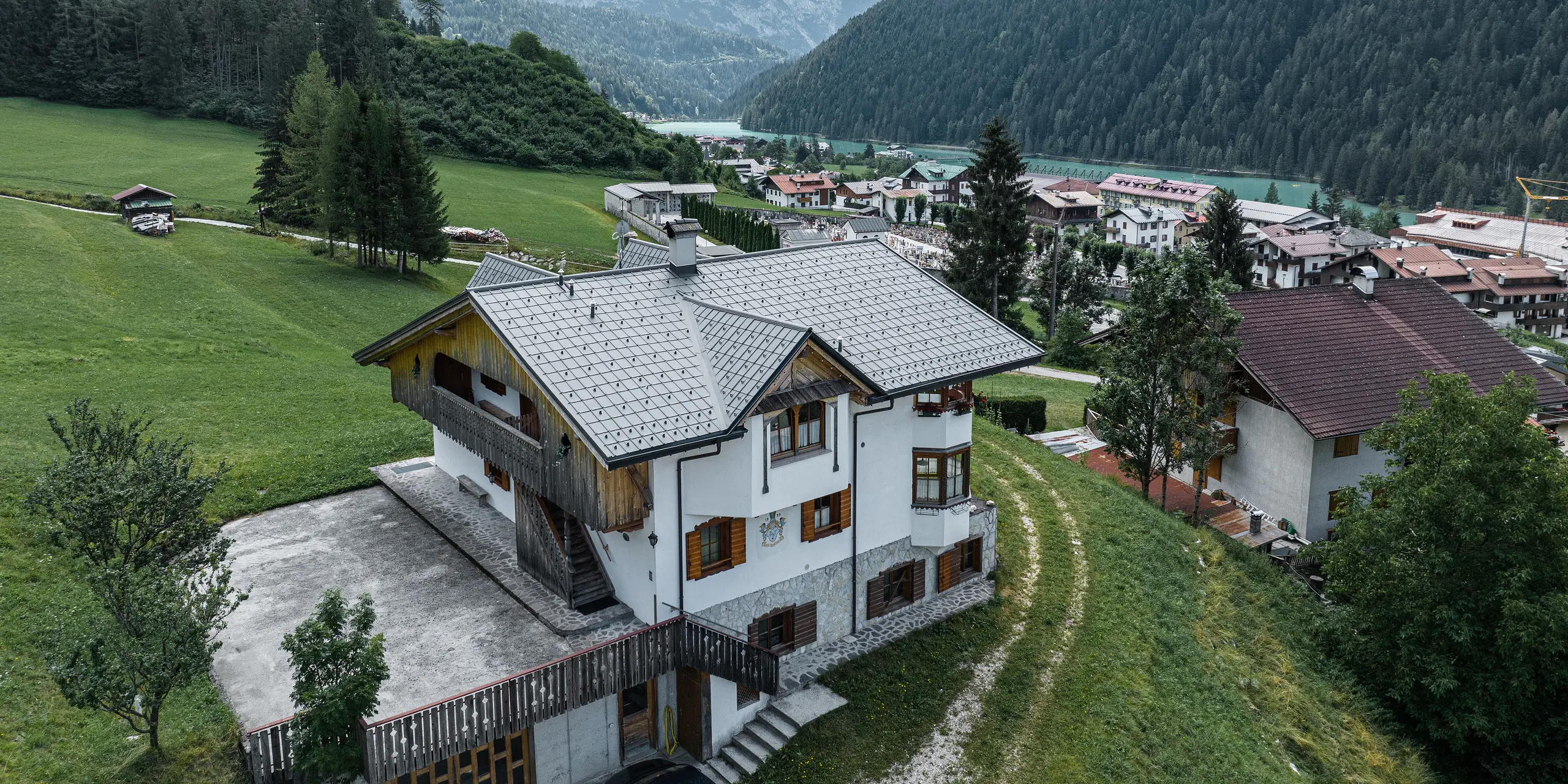 Luftaufnahme eines charmanten Bergchalets in Auronzo di Cadore, Italien, nach der Dachsanierung mit PREFA Aluminiumziegeln in Hellgrau. Das Chalet erhebt sich auf einer grünen Wiese mit einer beeindruckenden Bergkette im Hintergrund und zeigt ein malerisches Zusammenspiel von modernisierten Bauelementen und traditioneller Alpenarchitektur. Über dem Eingangsbereich ist eine Spitzgaube mit einem Holzgiebel zu sehen.