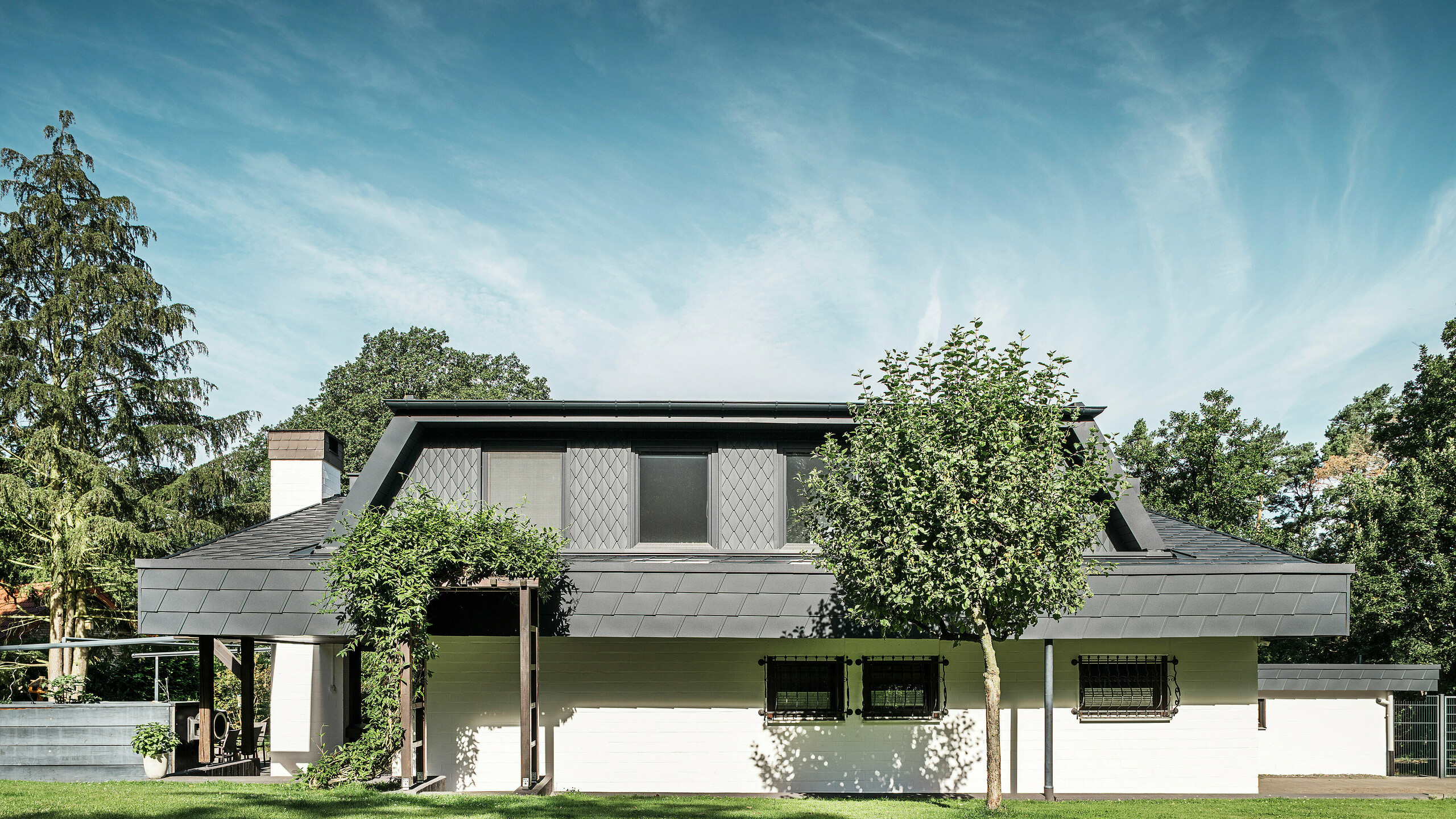 Widok z przodu na stronę ogrodową domu jednorodzinnego w Mechernich z dużą lukarną trapezową, pokrytą małymi dachówkami PREFA P.10 antracyt. Dach pokryty jest dachówkami łupkowymi PREFA DS.19 w tym samym odcieniu. Elewacja parteru jest pomalowana na biało, a zielona pergola dopełnia całości w części ogrodowej. Drzewa i krzewy otaczają widok i zapewniają harmonijne wkomponowanie w otoczenie.