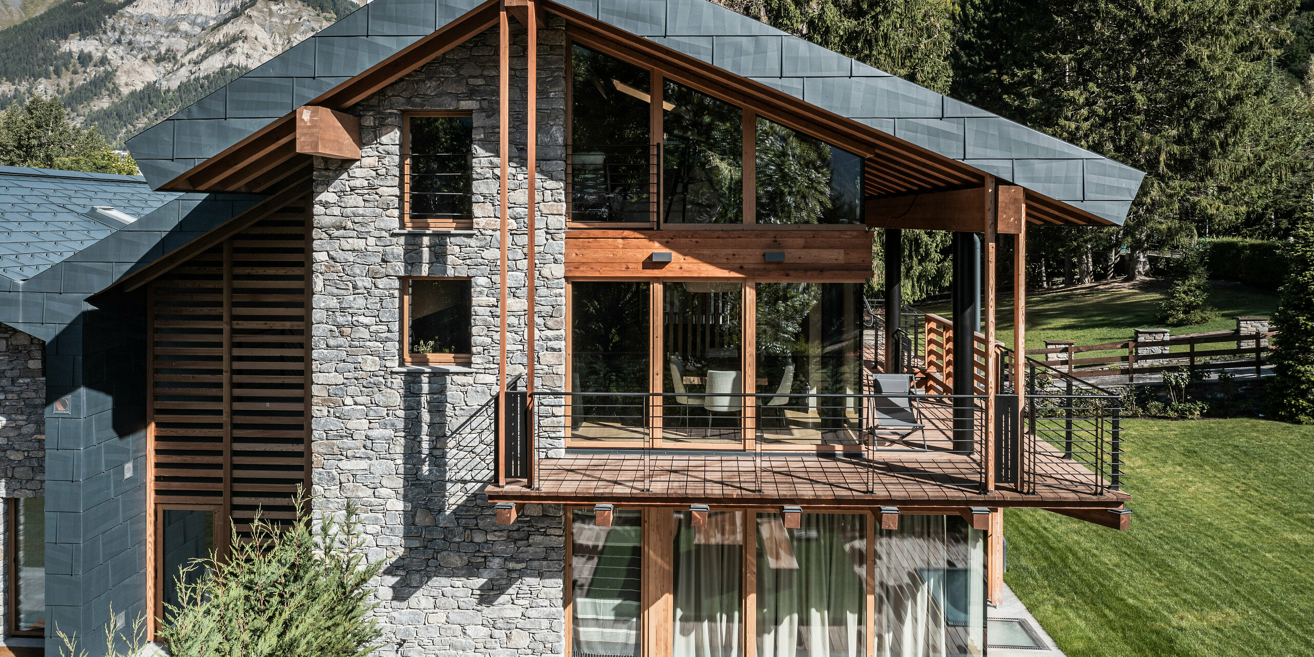Luxusní vila v Courmayeur s nápadnou kombinací dřeva, kamene a PREFA hliníku v podobě střešních a fasádních panelů FX.12 v barvě P.10 břidlicové. Strukturovaný fasádní obklad a velká čela oken nabízejí přirozenou estetiku. Velký krytý dřevěný balkon s výhledem na alpskou krajinu nabízí úchvatný výhled. Pečlivý design se hladce integruje s okolním horským pozadím a vytváří dramatické pozadí.