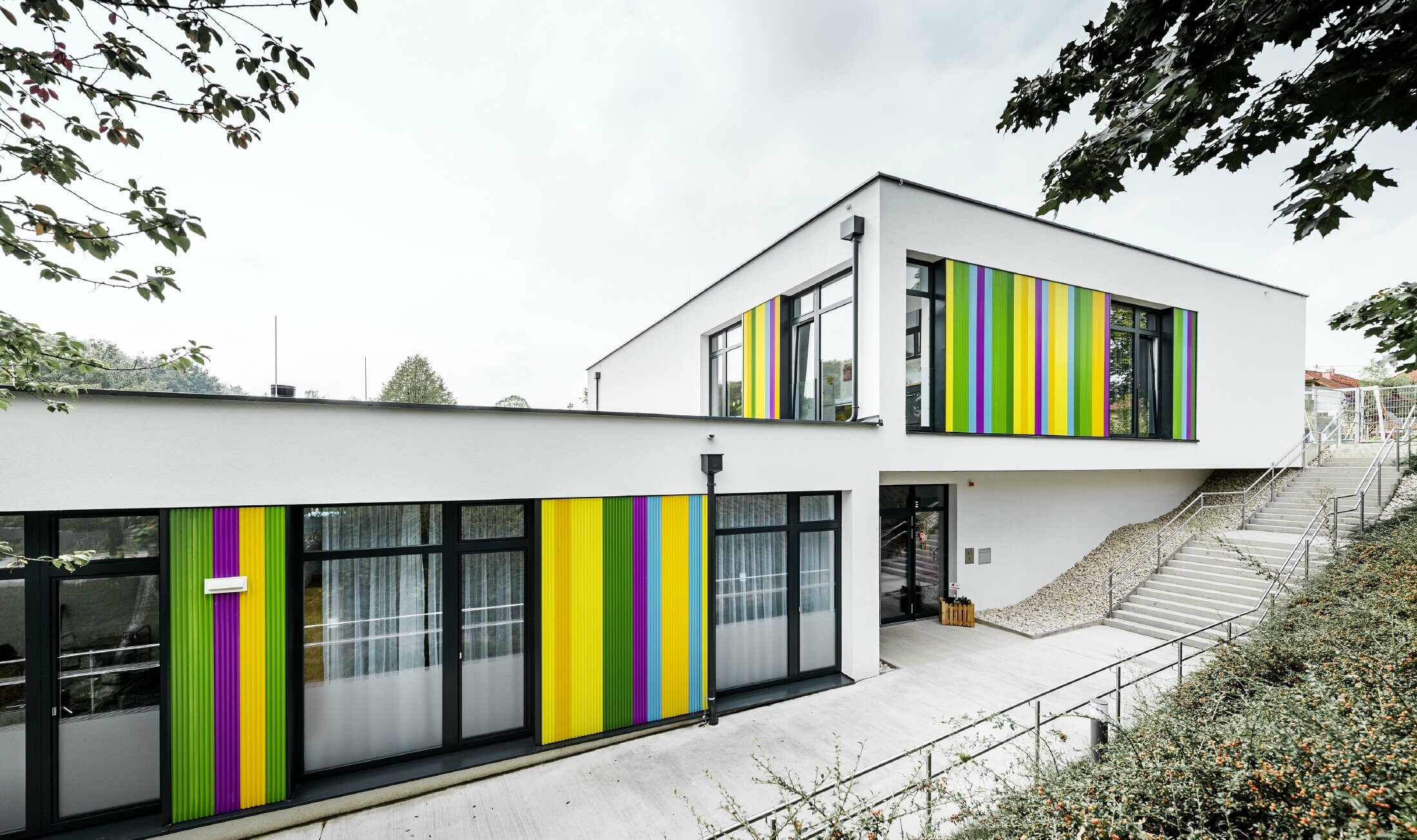 Kolorowe elementy elewacji PREFA przedszkola w Hargelsbergu. Budynek ma dach płaski i duże, dochodzące do ziemi okna.