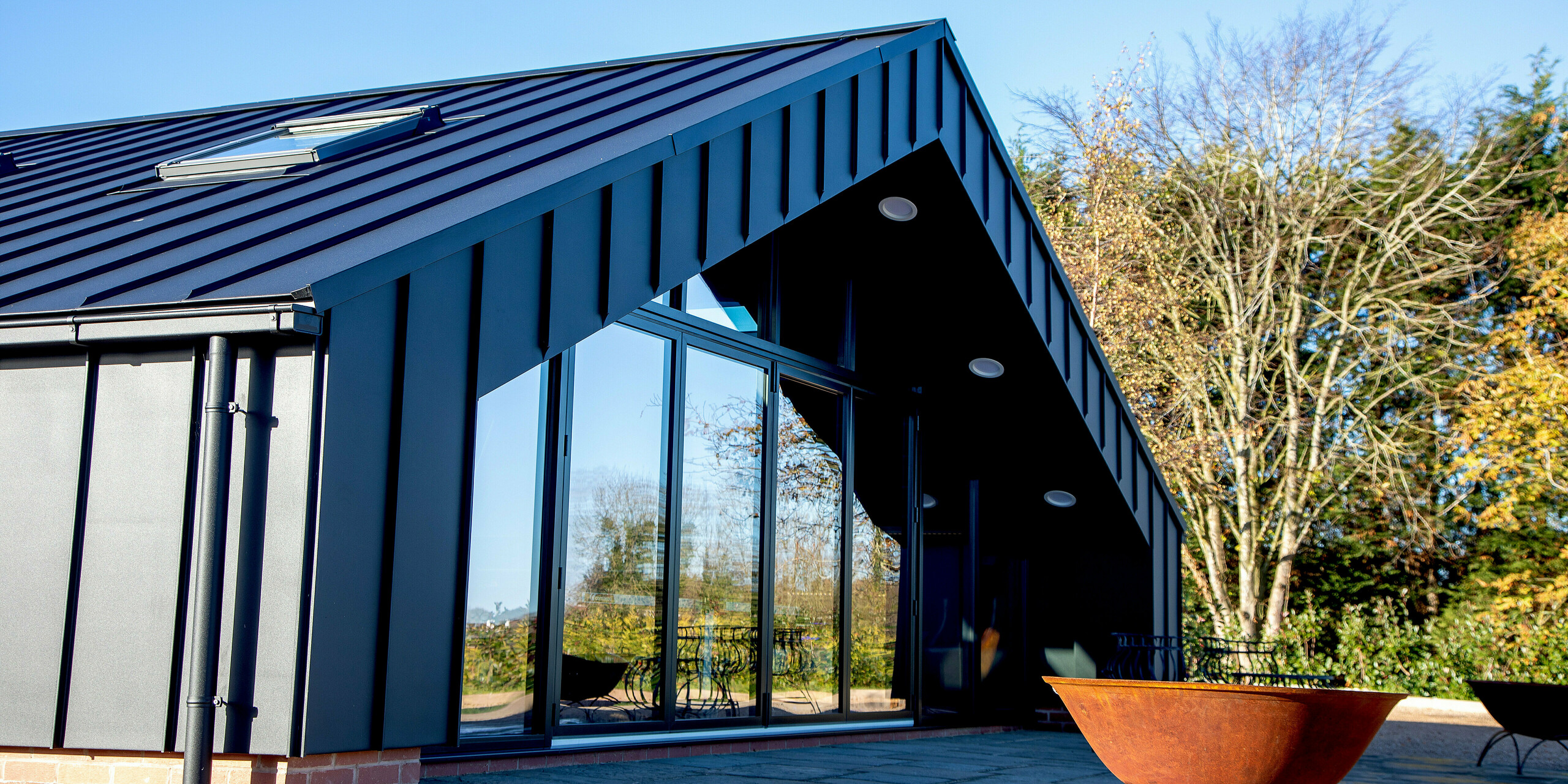 Froschperspektive auf das moderne Freizeitgebäude mit vollständig verkleideter PREFA Aluminiumfassade und PREFALZ Dachfläche in P.10 Schwarz. Die markanten Stehfalzlinien, die klare Dachgeometrie und die großzügige Glasfront erzeugen eine kraftvolle architektonische Präsenz. Die witterungsbeständige Aluminiumausführung garantiert höchste Stabilität und langlebigen Schutz im Außenbereich.