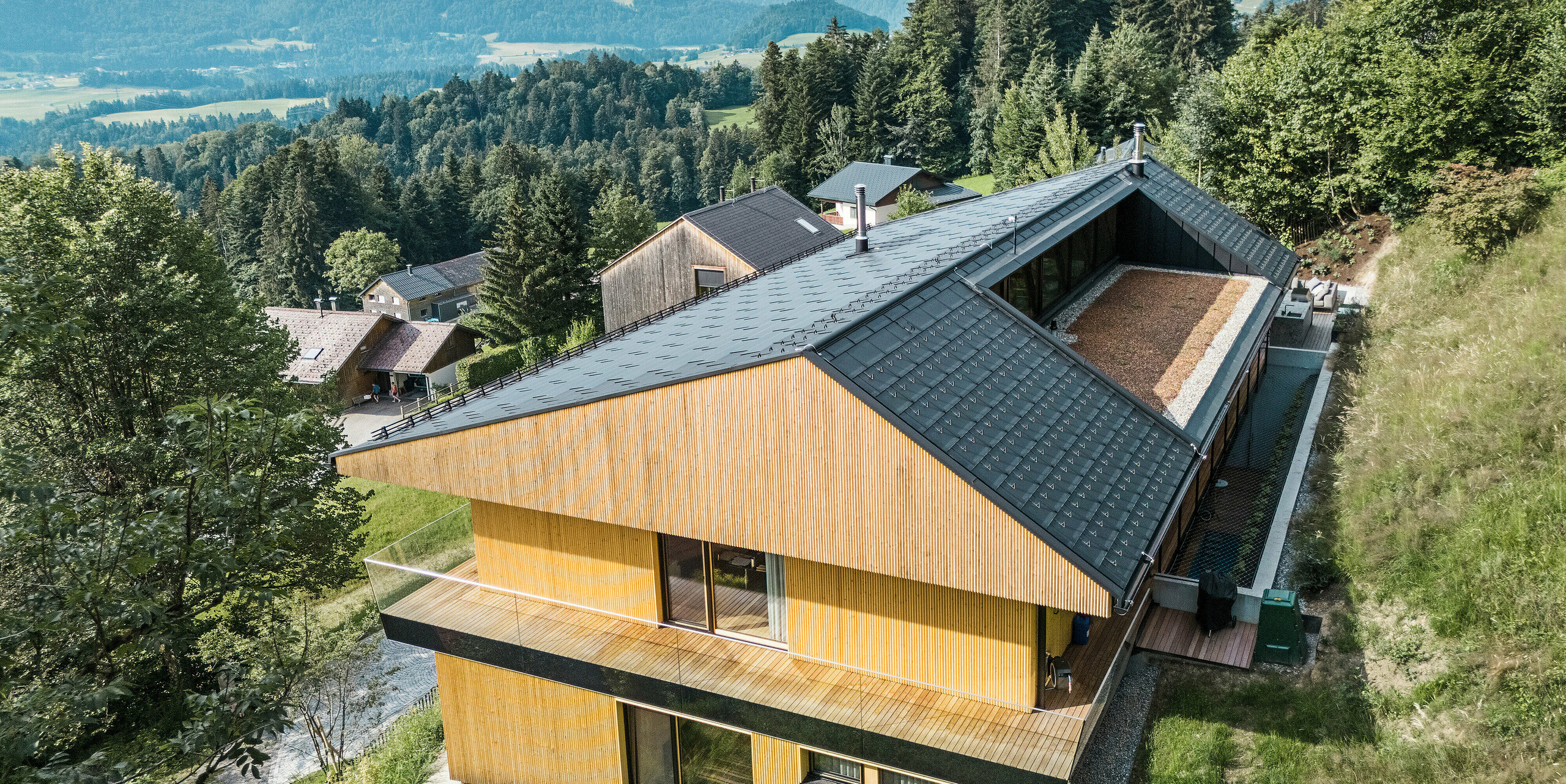 Blick auf die Holzfassade eines Einfamilienhauses in Egg, Vorarlberg. Das Gebäude wurde mit PREFA Solardachplatten und Dachplatten R.16 in P.10 Schwarz eingedeckt. Die moderne Dachkonstruktion aus Aluminium verfügt über eine harmonisch integrierte Dachloggia auf der Gebäuderückseite. Die natürliche Holzfassade fügt sich perfekt in die alpine Landschaft mit dem malerischen Bergpanorama im Hintergrund ein, während das robuste Aluminiumdach dauerhaften Schutz bietet.