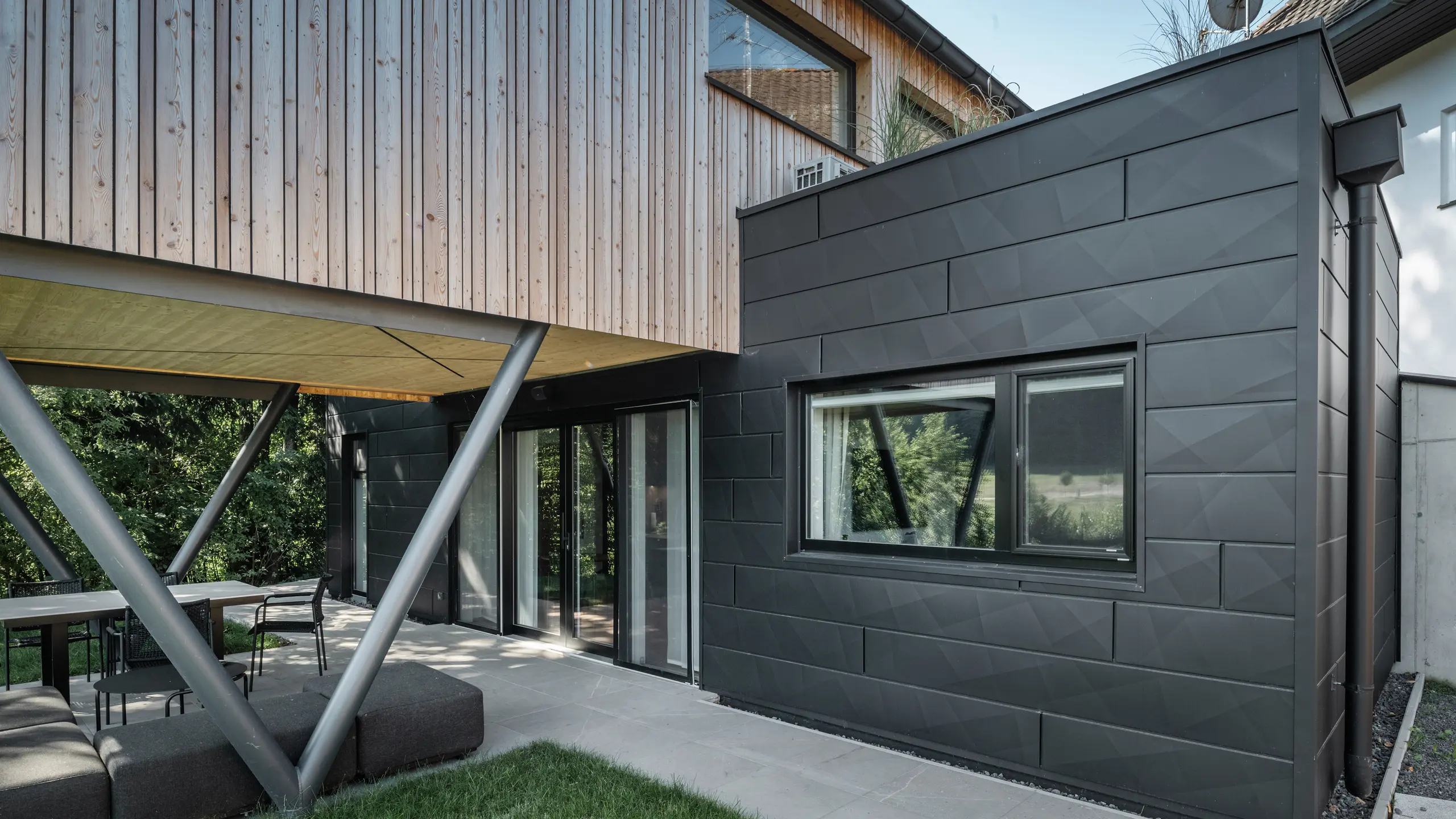 Atrakcja architektoniczna: „Woodstock Chalet 5” w St. Martin am Innkreis w Górnej Austrii. Nowoczesny obiekt przeznaczony na wesela wyróżnia się dzięki zastosowaniu paneli elewacyjnych PREFA Siding.X w kolorze antracytowym. Elementy aluminium tworzą silny kontrast z jasnymi panelami drewnianymi i metalowymi konstrukcjami nośnymi. Ciemne ramy okienne harmonijnie komponują się z wyglądem elewacji, nadając budynkowi elegancki wygląd. Zdjęcie to pokazuje estetyczną równowagę między naturalną fakturą drewna a industrialnym urokiem aluminiowej elewacji, która jest symbolem współczesnego budownictwa.