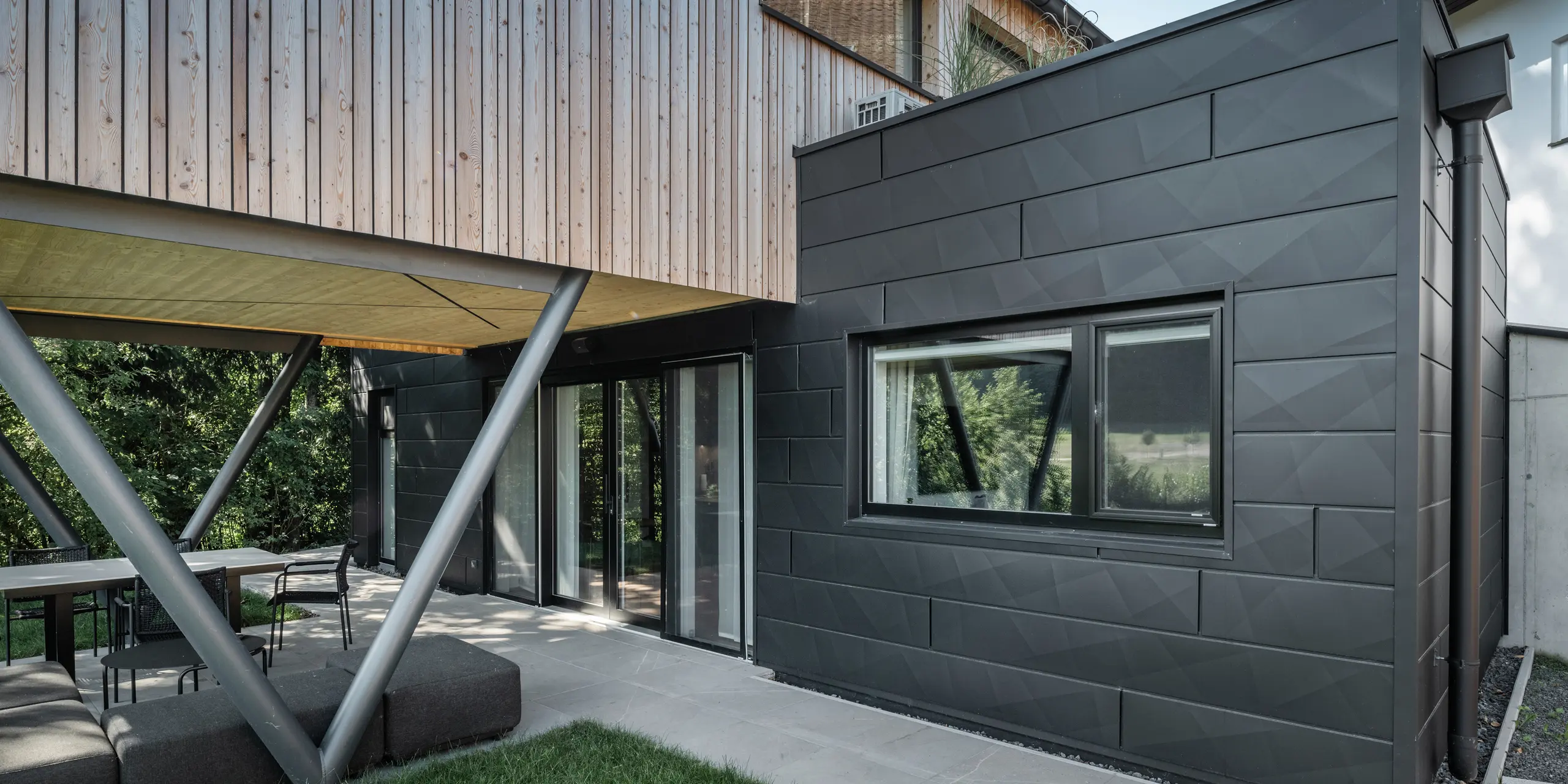 Atrakcja architektoniczna: „Woodstock Chalet 5” w St. Martin am Innkreis w Górnej Austrii. Nowoczesny obiekt przeznaczony na wesela wyróżnia się dzięki zastosowaniu paneli elewacyjnych PREFA Siding.X w kolorze antracytowym. Elementy aluminium tworzą silny kontrast z jasnymi panelami drewnianymi i metalowymi konstrukcjami nośnymi. Ciemne ramy okienne harmonijnie komponują się z wyglądem elewacji, nadając budynkowi elegancki wygląd. Zdjęcie to pokazuje estetyczną równowagę między naturalną fakturą drewna a industrialnym urokiem aluminiowej elewacji, która jest symbolem współczesnego budownictwa.