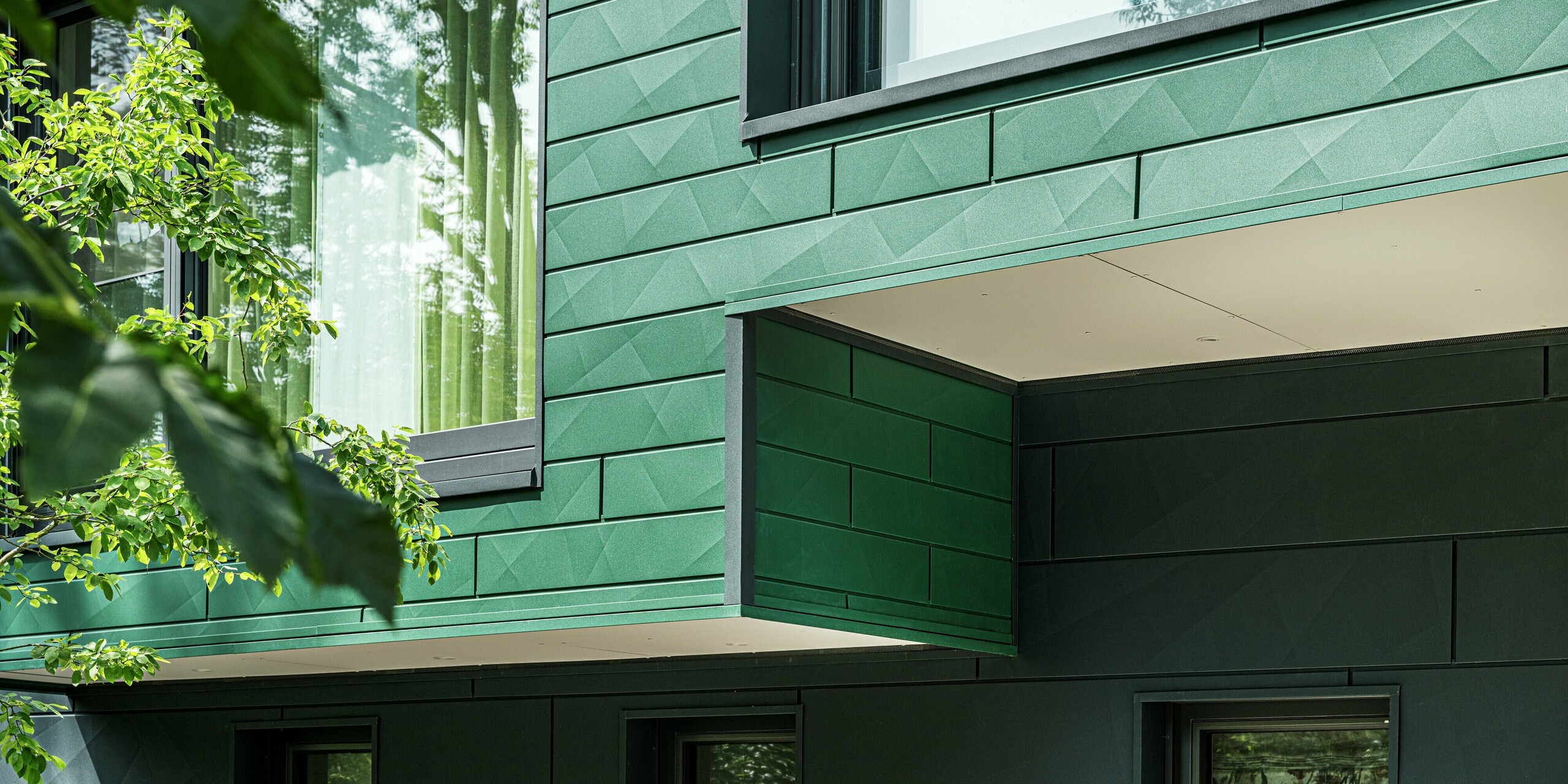 Das Detailbild zeigt die markante Fassade des in Türkheim gelegenen Einfamilienhauses, das mit PREFA Siding X-Paneelen in den Farben Moosgrün und Anthrazit verkleidet wurde. Die Längs- und Querkantungen der Paneele schaffen eine interessante Struktur und verleihen der Fassade eine  dynamische Optik. Das grüne Farbschema harmoniert perfekt mit der Umgebung und den Bäumen im Vordergrund. Zudem überzeugt die Aluminiumfassade durch ihre Langlebigkeit und Witterungsbeständigkeit.