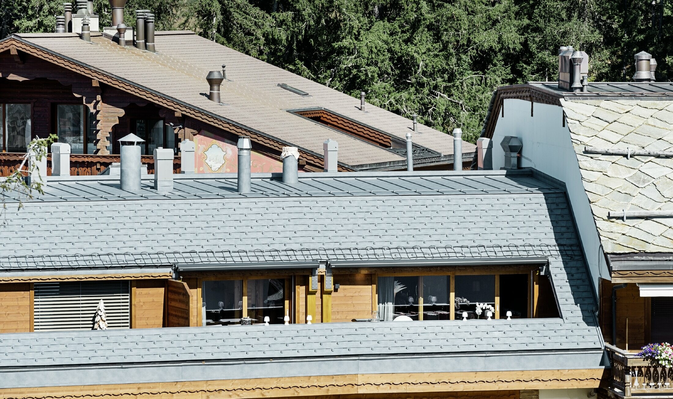 Budynek wielorodzinny w Crans-Montana z górami w tle i elewacją z fantazyjnymi elementami drewnianymi skomponowanymi z aluminiową dachówką łupkową PREFA w kolorze kamiennym szarym