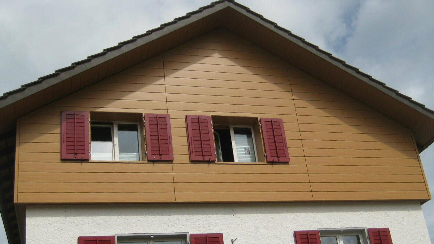 Elewacja budynku w kolorze drewna, z sidingami PREFA w układzie poziomym, okna z czerwonymi okiennicami