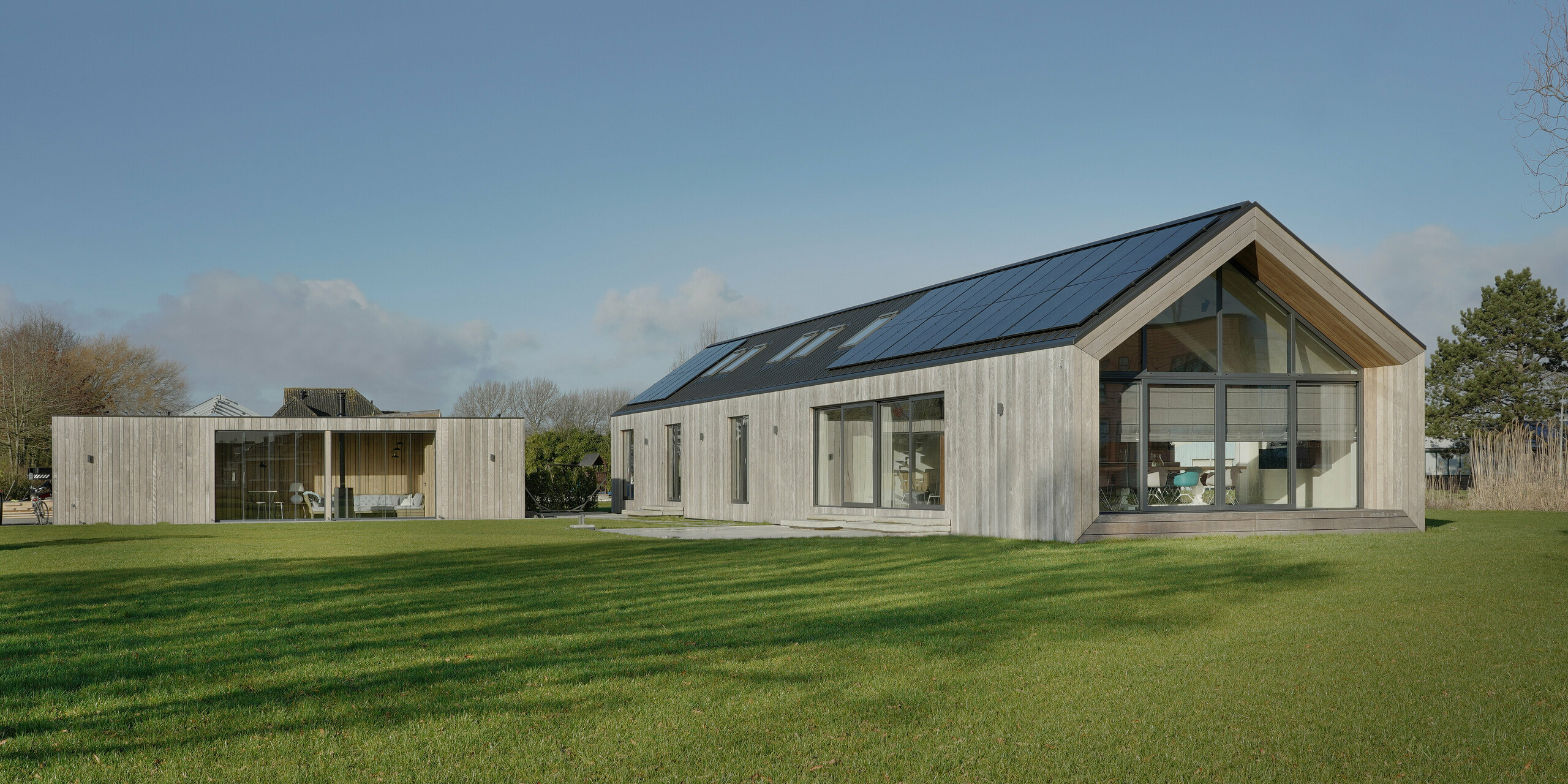 Weitwinkelaufnahme eines modernen Einfamilienhauses in Uithoorn, Niederlande, mit PREFALZ Winkelstehfalz-Eindeckung in Schwarzgrau und integrierter Photovoltaik-Anlage. Das Bild zeigt das gesamte Anwesen mit großzügigem Garten und einer Kombination aus großen Glasfronten und natürlichen Holzelementen an der Fassade. Die klare Linienführung und die hochwertigen, wetterbeständigen Materialien betonen die Nachhaltigkeit und das moderne Design des Hauses. Der einstöckige Bungalow wurde für die Wohnbedürfnisse einer Großfamilie erbaut.