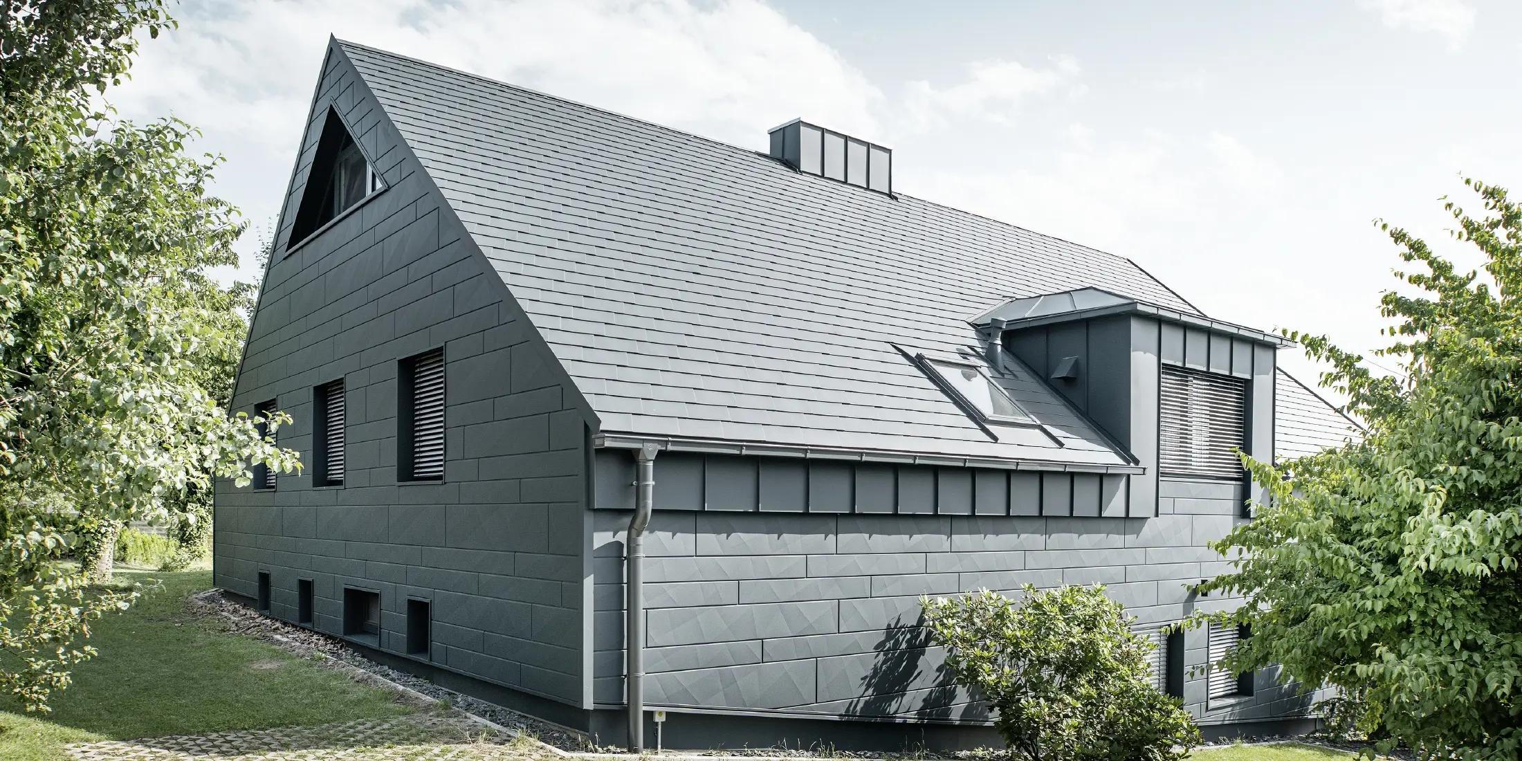 Duży dom jednorodzinny z dachem dwuspadowym i wieloma oknami. Cały dom został pokryty aluminiową okładziną w kolorze antracytowym, na dachu zastosowano aluminiowe gonty dachowe PREFA, a na elewacji siding.X w kolorze antracytowym.