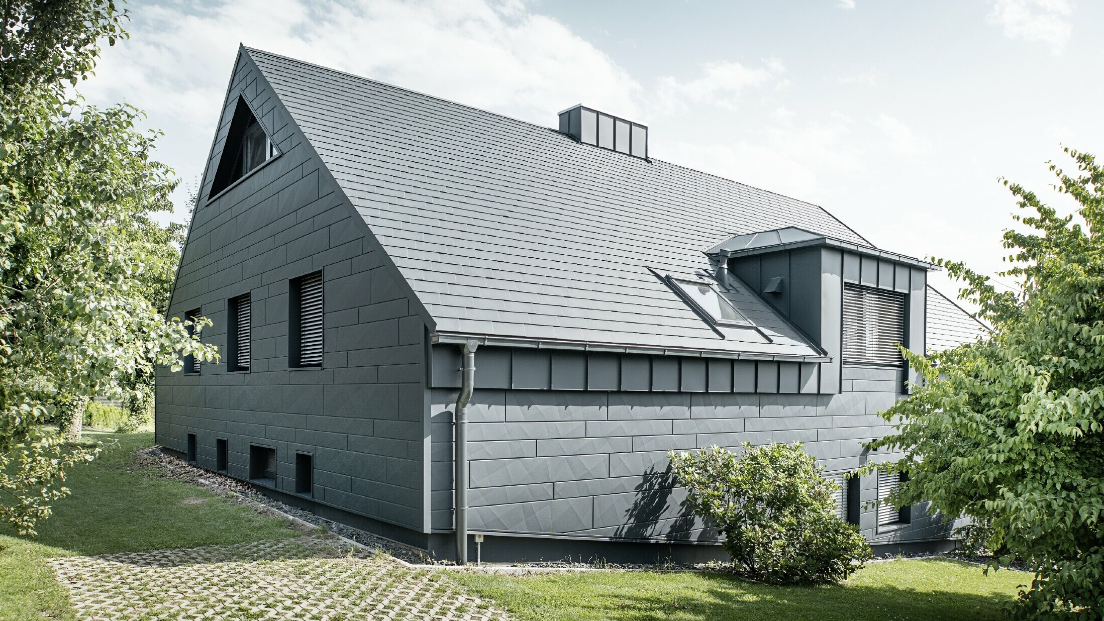 Duży dom jednorodzinny z dachem dwuspadowym i wieloma oknami. Cały dom został pokryty aluminiową okładziną w kolorze antracytowym, na dachu zastosowano aluminiowe gonty dachowe PREFA, a na elewacji siding.X w kolorze antracytowym.