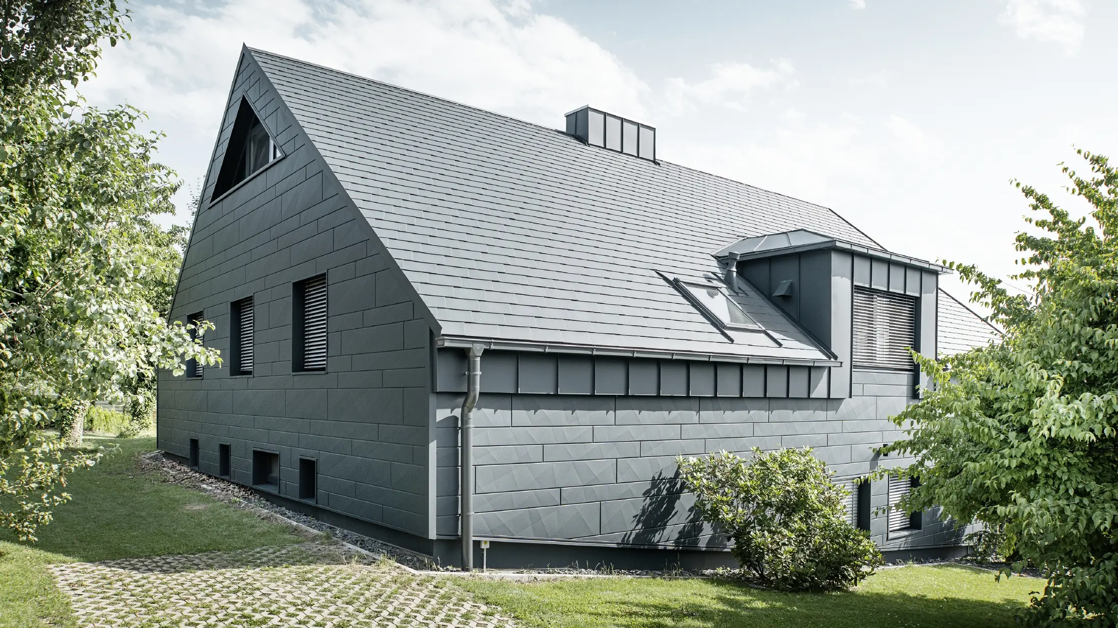 Duży dom jednorodzinny z dachem dwuspadowym i wieloma oknami. Cały dom został pokryty aluminiową okładziną w kolorze antracytowym, na dachu zastosowano aluminiowe gonty dachowe PREFA, a na elewacji siding.X w kolorze antracytowym.