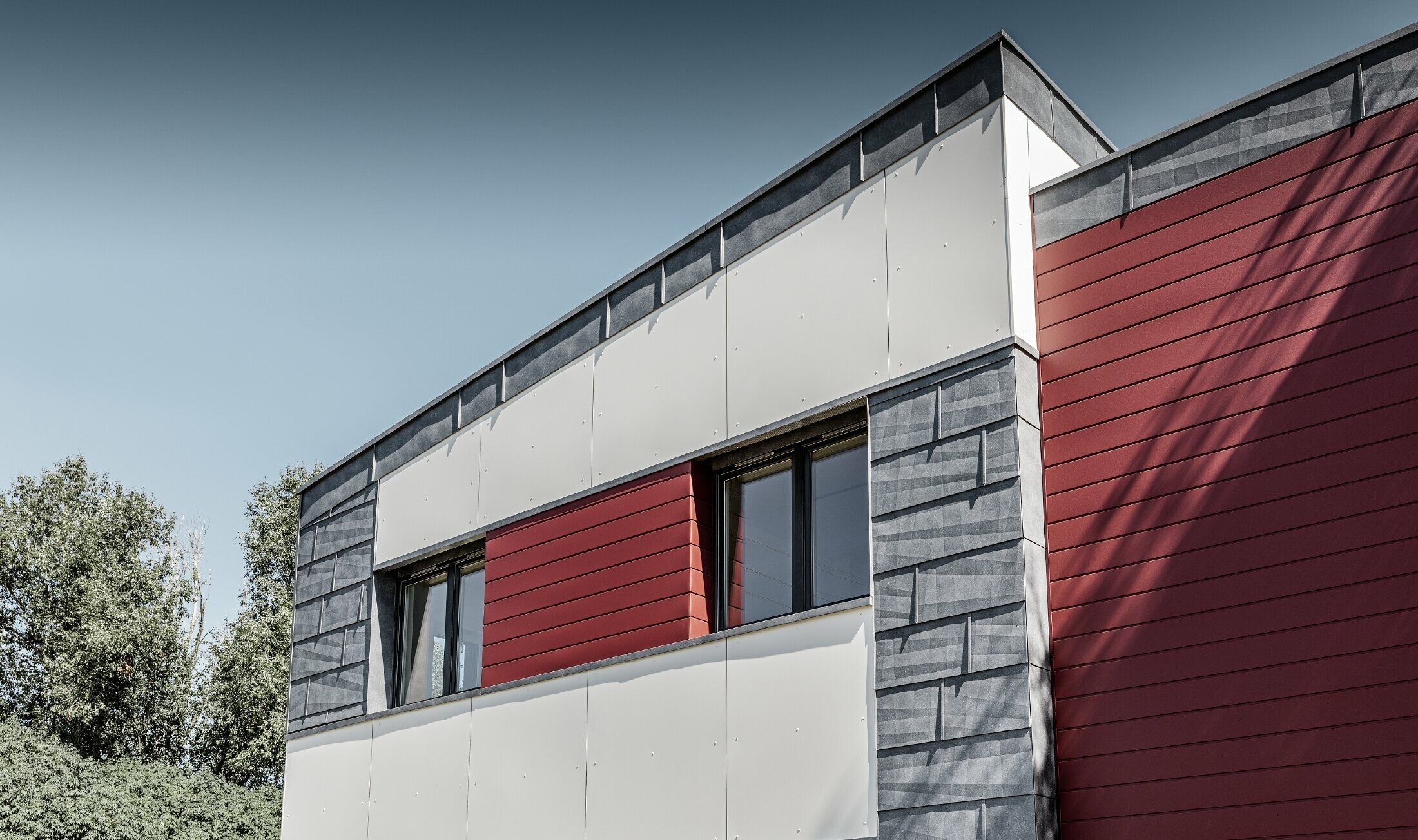 Nowoczesny budynek biurowy z dzieloną elewacją w kolorach czerwonym, kamiennym szarym i białym, wykonaną z aluminiowego sidingu, paneli elewacyjnych FX.12 i aluminiowej płyty kompozytowej PREFA