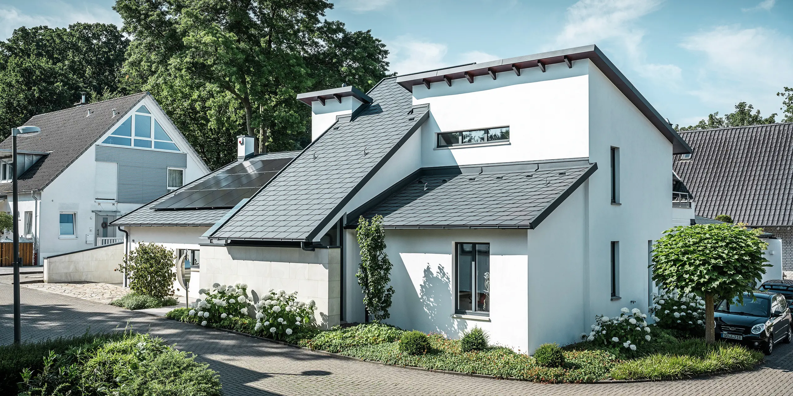 Nowoczesny dom jednorodzinny z białą elewacją pokrytą tynkiem i ciemnym dachem skośnym PREFA z aluminium. Na dachu widoczne są zintegrowane moduły fotowoltaiczne i aluminiowe odwodnienie dachu w kolorze antracyt. Budynek położony jest na zadbanym osiedlu mieszkaniowym z zadbanymi ogrodami przed domami, brukowanym podjazdem i otaczającymi domami jednorodzinnymi.
