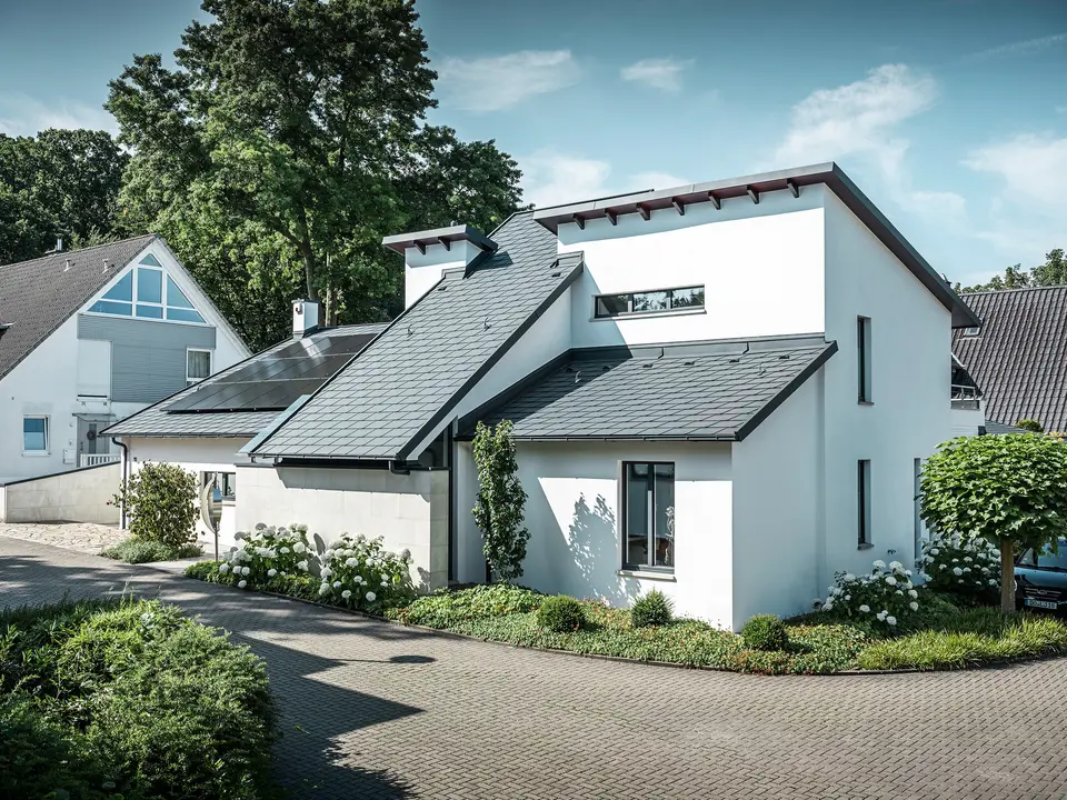 Nowoczesny dom jednorodzinny z białą elewacją pokrytą tynkiem i ciemnym dachem skośnym PREFA z aluminium. Na dachu widoczne są zintegrowane moduły fotowoltaiczne i aluminiowe odwodnienie dachu w kolorze antracyt. Budynek położony jest na zadbanym osiedlu mieszkaniowym z zadbanymi ogrodami przed domami, brukowanym podjazdem i otaczającymi domami jednorodzinnymi.