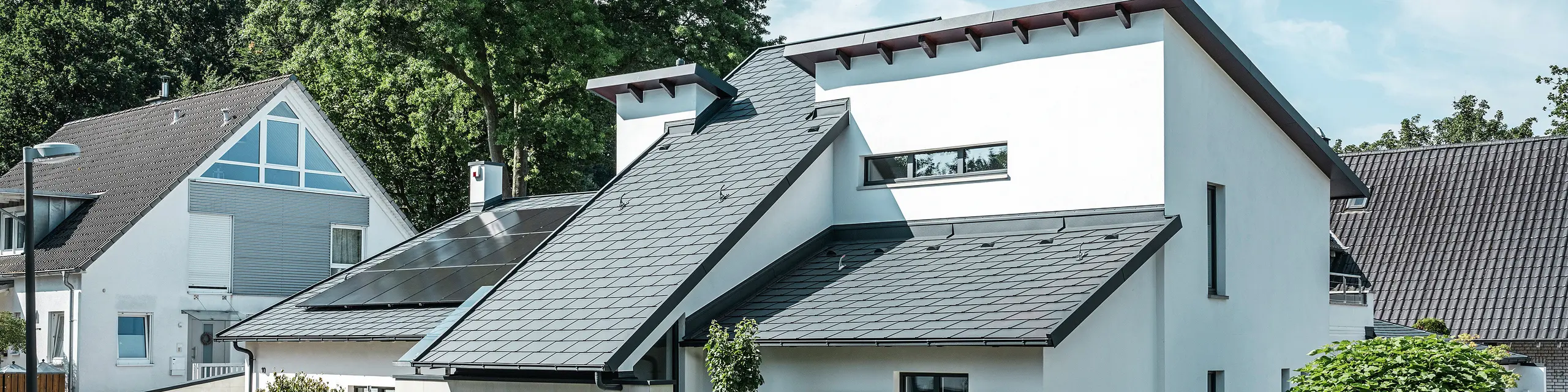 Nowoczesny dom jednorodzinny z białą elewacją pokrytą tynkiem i ciemnym dachem skośnym PREFA z aluminium. Na dachu widoczne są zintegrowane moduły fotowoltaiczne i aluminiowe odwodnienie dachu w kolorze antracyt. Budynek położony jest na zadbanym osiedlu mieszkaniowym z zadbanymi ogrodami przed domami, brukowanym podjazdem i otaczającymi domami jednorodzinnymi.