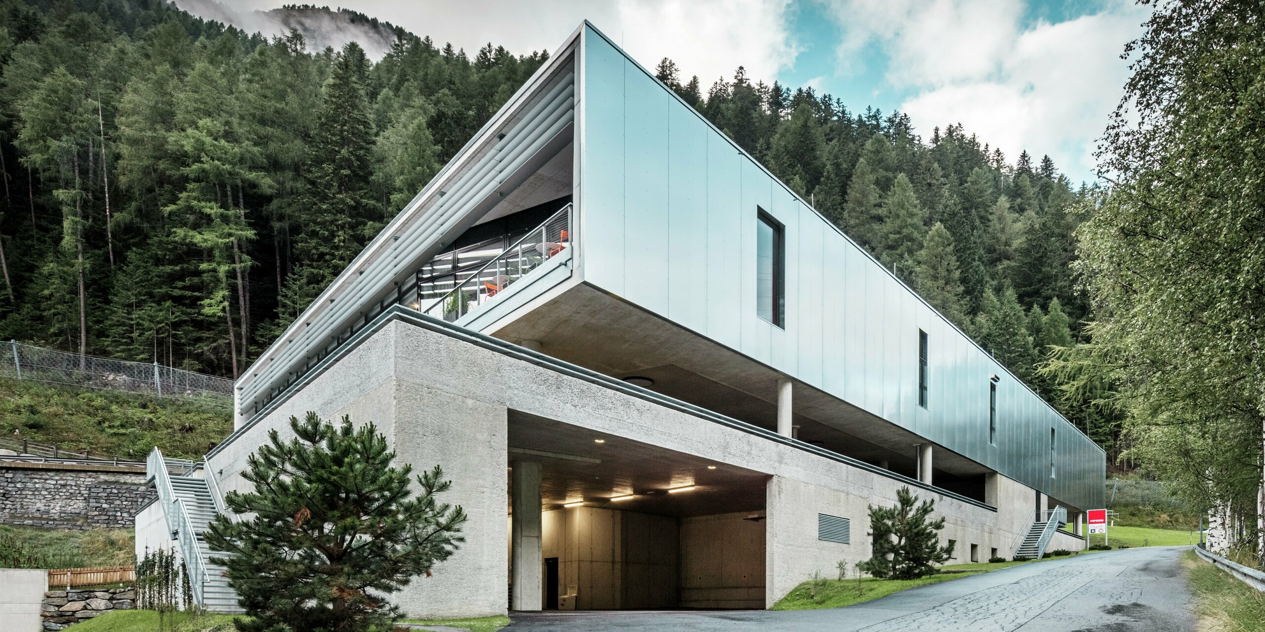 Rückansicht des MPREIS-Marktes in Sölden, Tirol, mit reflektierenden PREFA Aluminium Verbundplatten in der Sonderfarbe Reflective Natural. Die moderne Architektur fügt sich in die bewaldete Alpenlandschaft ein und verbindet Funktionalität mit Ästhetik. Das Parkdeck und die Zufahrtsstraße sind eine praktische Lösung für das Bauen in Hanglage.