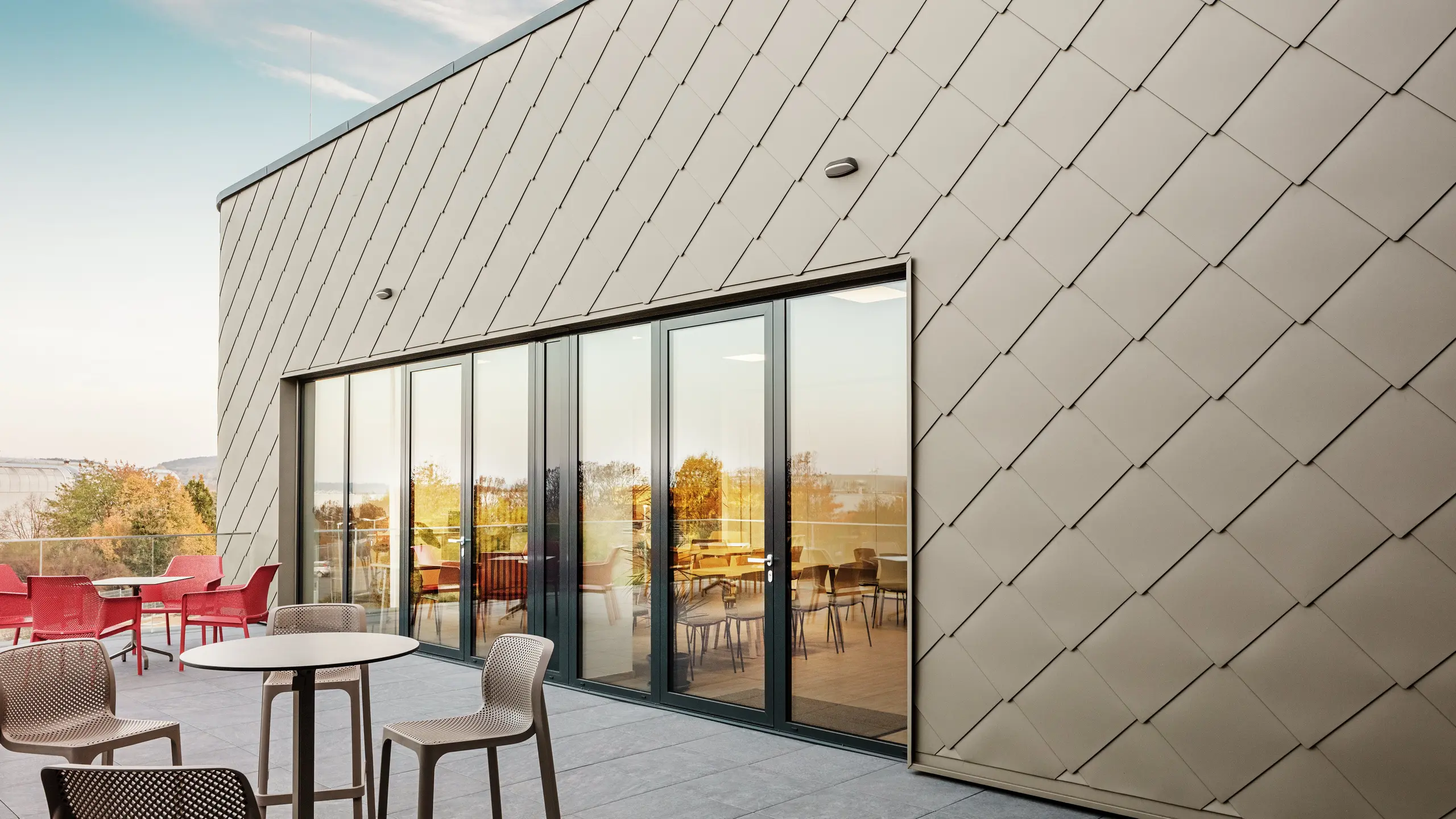 [Translate to Polski:] Die Fassade des neuen PREFA Standortes in Törökbálint besticht durch die eleganten PREFA Wandrauten 44 × 44 in Bronze. Die präzise verlegte Aluminiumfassade sorgt für ein modernes, dynamisches Erscheinungsbild und bietet gleichzeitig maximalen Schutz vor Witterungseinflüssen. Die großzügige Glasfront verbindet Innen- und Außenbereich und schafft auf der großzügigen Terrasse eine helle und einladende Atmosphäre.