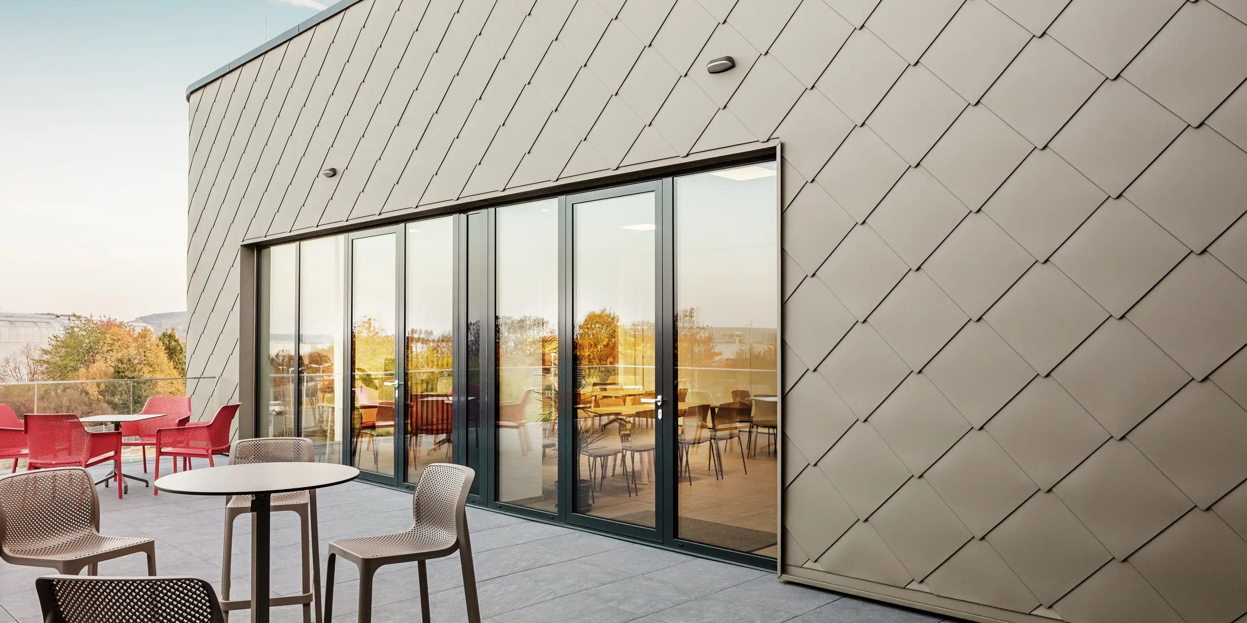 [Translate to Polski:] Die Fassade des neuen PREFA Standortes in Törökbálint besticht durch die eleganten PREFA Wandrauten 44 × 44 in Bronze. Die präzise verlegte Aluminiumfassade sorgt für ein modernes, dynamisches Erscheinungsbild und bietet gleichzeitig maximalen Schutz vor Witterungseinflüssen. Die großzügige Glasfront verbindet Innen- und Außenbereich und schafft auf der großzügigen Terrasse eine helle und einladende Atmosphäre.