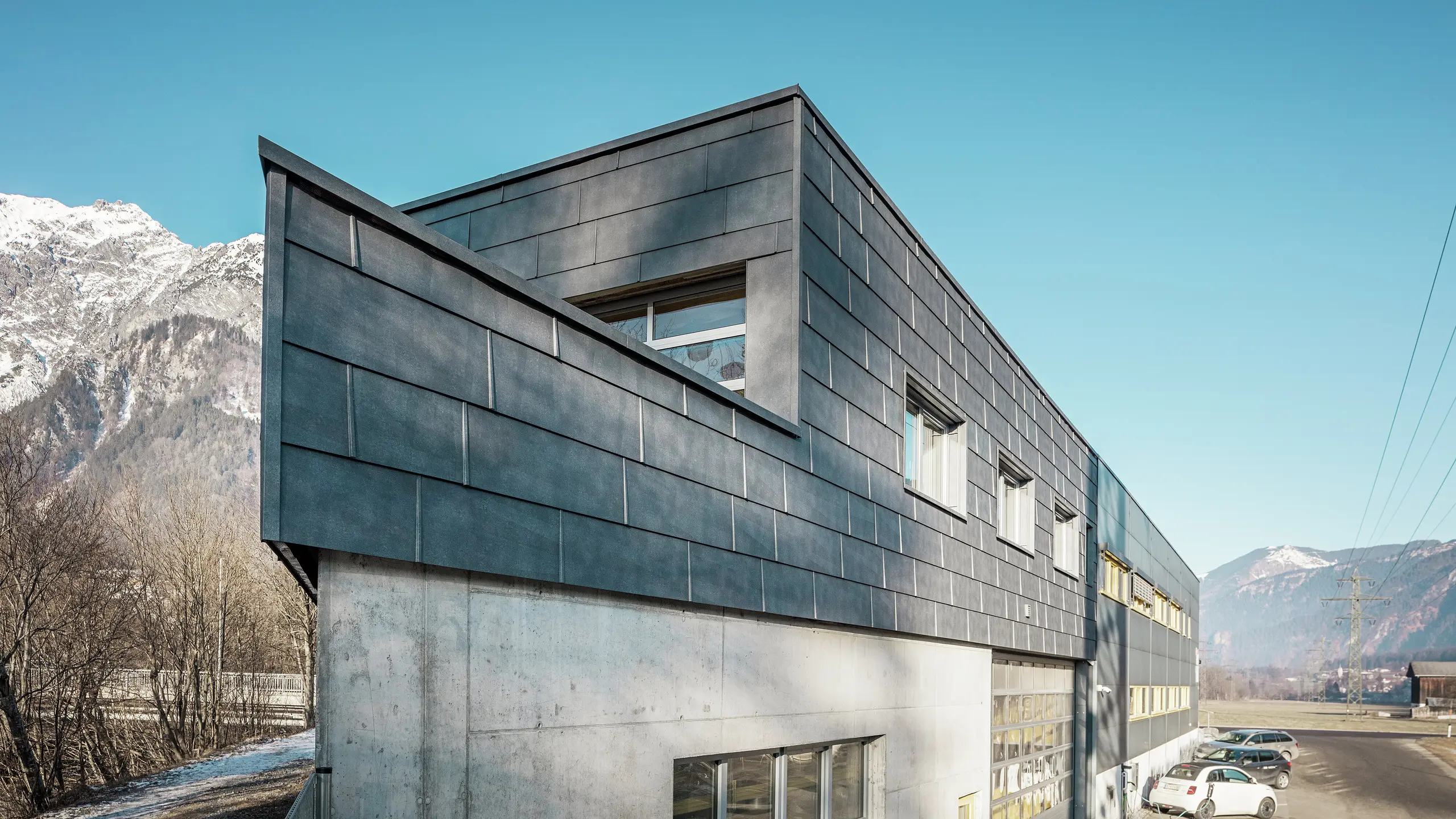 Przód budynku firmy blacharskiej RÜMA w Vorarlbergu. Budynek ma charakterystyczną elewację z szarych panelów elewacyjnych PREFA FX.12 z aluminium i widoczną betonową podstawą. Jest on położony w alpejskim krajobrazie z górami i błękitnym niebem. Na parkingu przed budynkiem widać ładujący się samochód elektryczny, co podkreśla zrównoważony charakter działalności firmy.