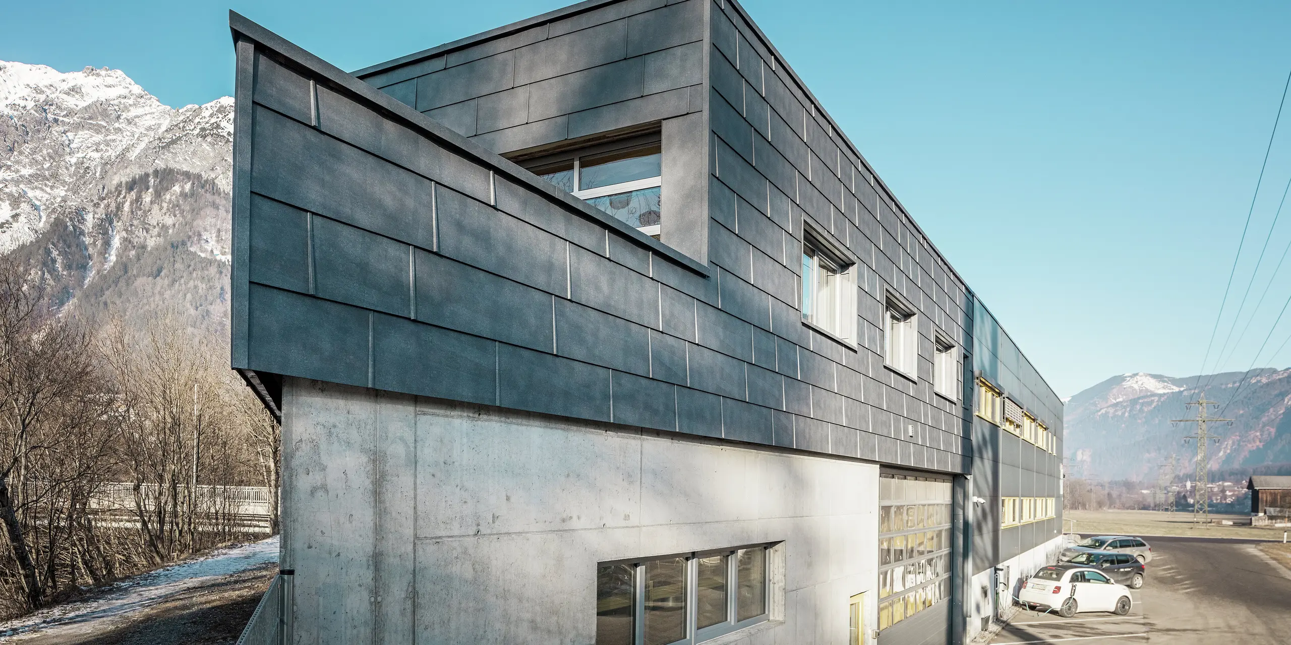 Przód budynku firmy blacharskiej RÜMA w Vorarlbergu. Budynek ma charakterystyczną elewację z szarych panelów elewacyjnych PREFA FX.12 z aluminium i widoczną betonową podstawą. Jest on położony w alpejskim krajobrazie z górami i błękitnym niebem. Na parkingu przed budynkiem widać ładujący się samochód elektryczny, co podkreśla zrównoważony charakter działalności firmy.