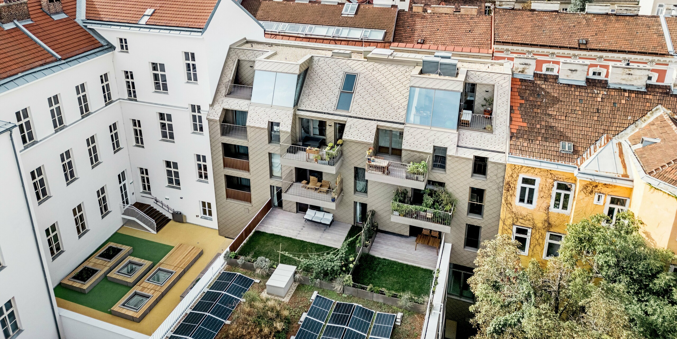 Die Vogelperspektive zeigt eine beeindruckende Aussicht auf die Erweiterung eines Wohngebäudes in Wien, bei dem PREFA Rauten 29×29 in Bronze auf der Dach- und Fassadengestaltung verwendet wurden. Die modernen Rauten harmonieren perfekt mit der traditionellen Architektur der Umgebung und schaffen eine attraktive visuelle Verbindung zwischen Alt und Neu. Im Innenhof ist ein durchdachtes Design mit Solarzellen, grünen Flächen und Holzterrassen zu sehen, das den nachhaltigen Ansatz der Wohnanlage unterstreicht. Die lebendige Mischung aus urbanem Raum und Natur wird durch die Dachterrassen und Balkone ergänzt, die den Bewohnern einen exklusiven Blick über die Wiener Dächer bieten.