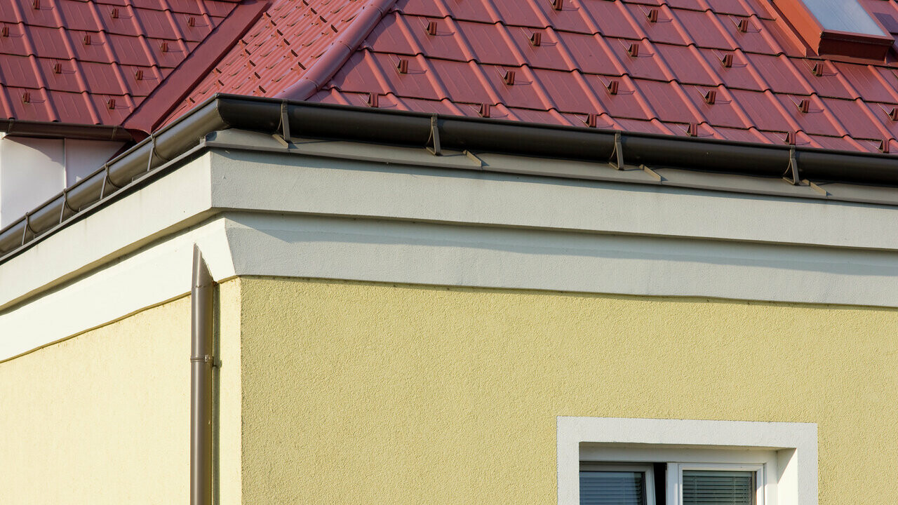Narożnik domu z żółtą elewacją tynkową i brązową rynnę PREFA (rynna dachowa) z czerwonym dachem z płyt dachowych PREFA