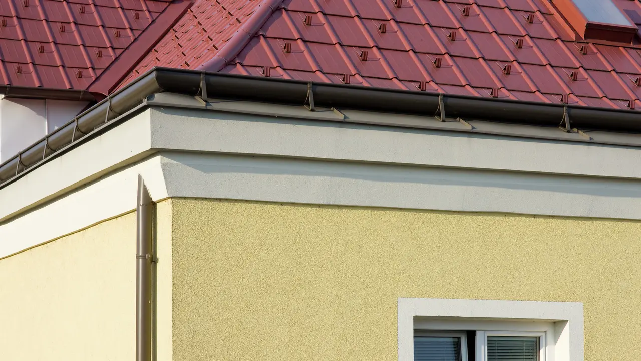 Narożnik domu z żółtą elewacją tynkową i brązową rynnę PREFA (rynna dachowa) z czerwonym dachem z płyt dachowych PREFA