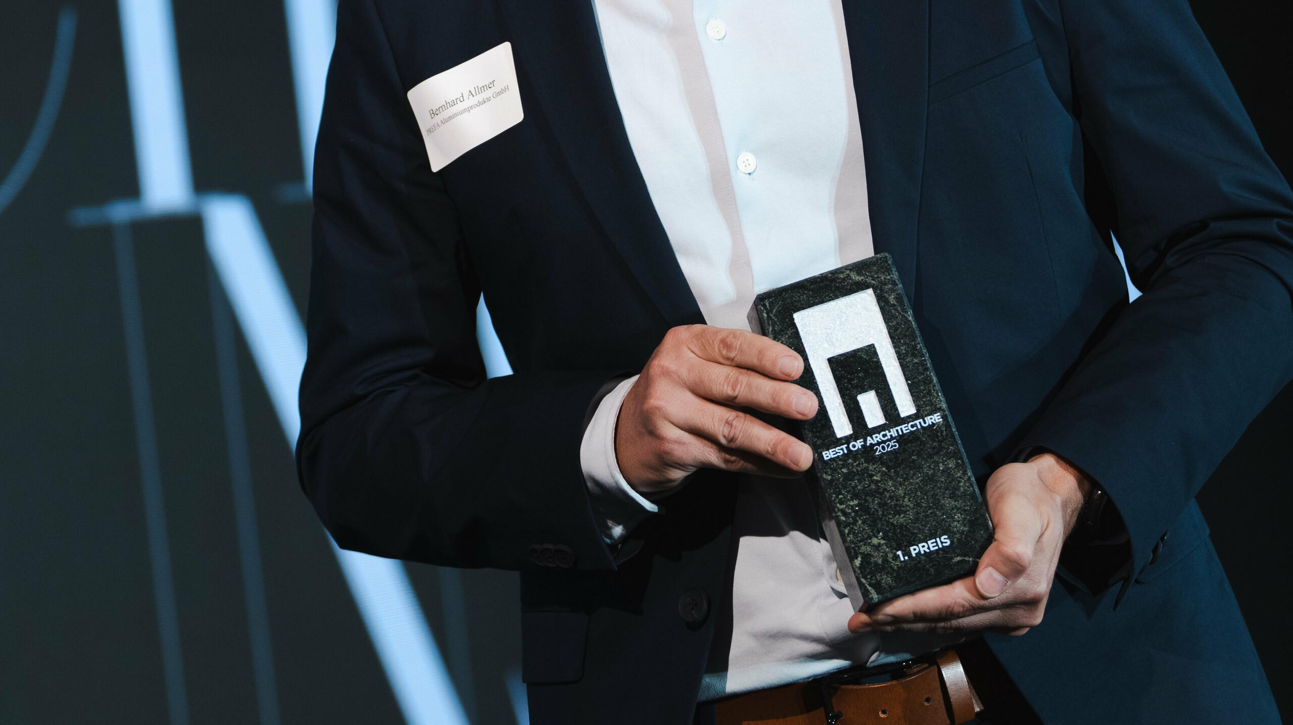 Ceremonia wręczenia nagród w renomowanym konkursie Callwey „Budynek mieszkalny roku” 2025: na pierwszym planie elegancka postać w ciemnym garniturze i białej koszuli dumnie prezentuje nagrodę „Architects' Choice”. Osobą tą jest Bernhard Allmer, odpowiedzialny za produkty solarne PREFA. Cena składa się z eleganckiej ciemnej płyty kamiennej z nowoczesnym logo i wyraźną typografią. W tle znajduje się stylowa scena z dużym banerem i oświetleniem akcentującym, które podkreśla uroczysty charakter wydarzenia. Scena odzwierciedla innowacyjność i doskonałość w architekturze.