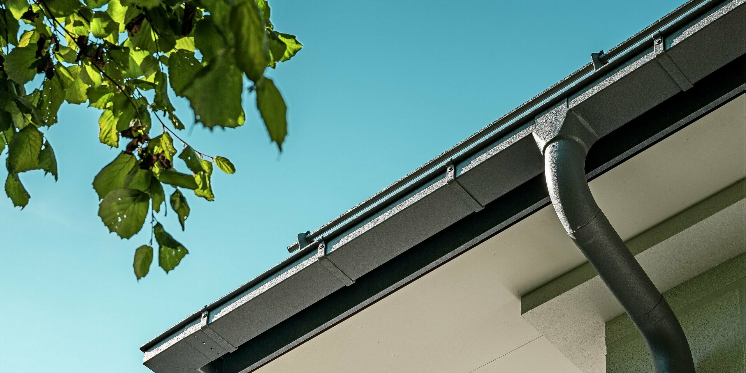 Detailaufnahme der PREFA Dachentwässerung mit Kastenrinne und Ablaufrohr in der eleganten Farbe P.10 Steingrau. Das hochwertige Aluminium-System sorgt für eine effiziente Wasserableitung und passt sich nahtlos in das moderne Design des Mehrfamilienhauses ein. Die präzise Verarbeitung der Kastenrinne und das zeitgemäße Erscheinungsbild unterstreichen die ästhetische und funktionale Qualität des Daches. Die klare Linienführung und das ansprechende Material machen dieses Dachentwässerungssystem zu einem wichtigen Designelement des Gebäudes.